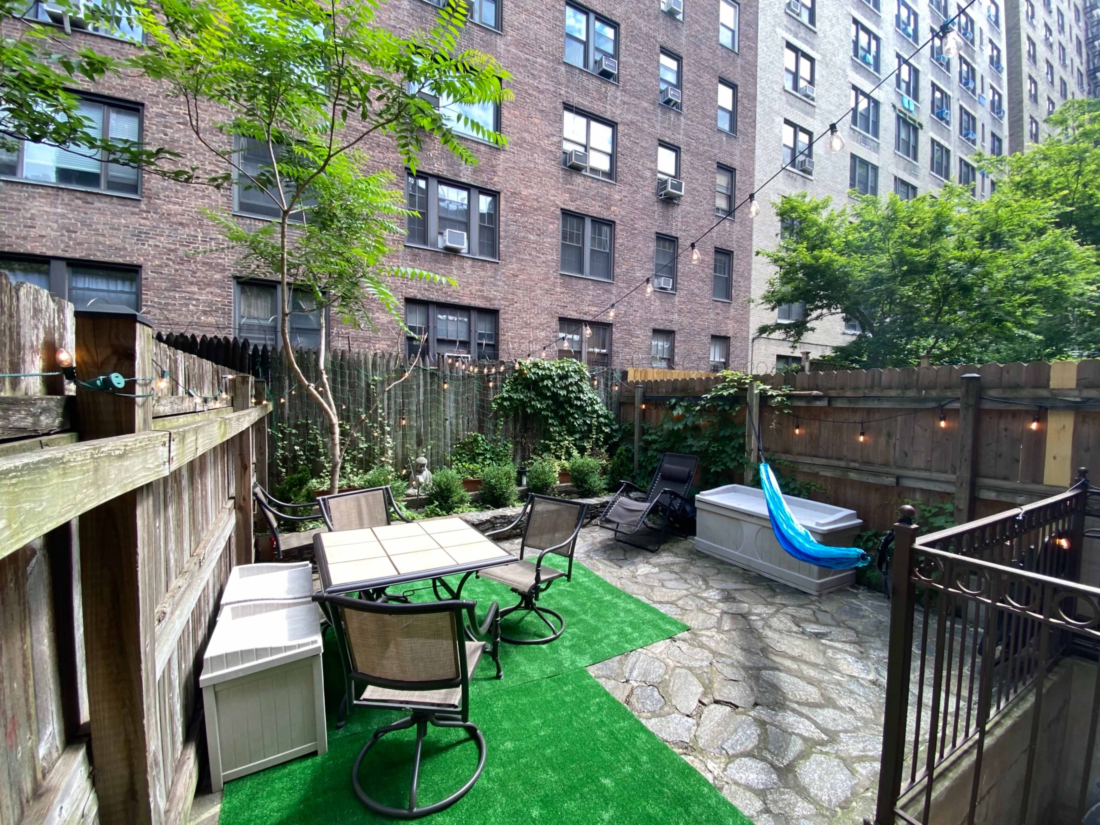 The image shows a small backyard patio with a table and chairs, a hot tub, and greenery, surrounded by tall apartment buildings.