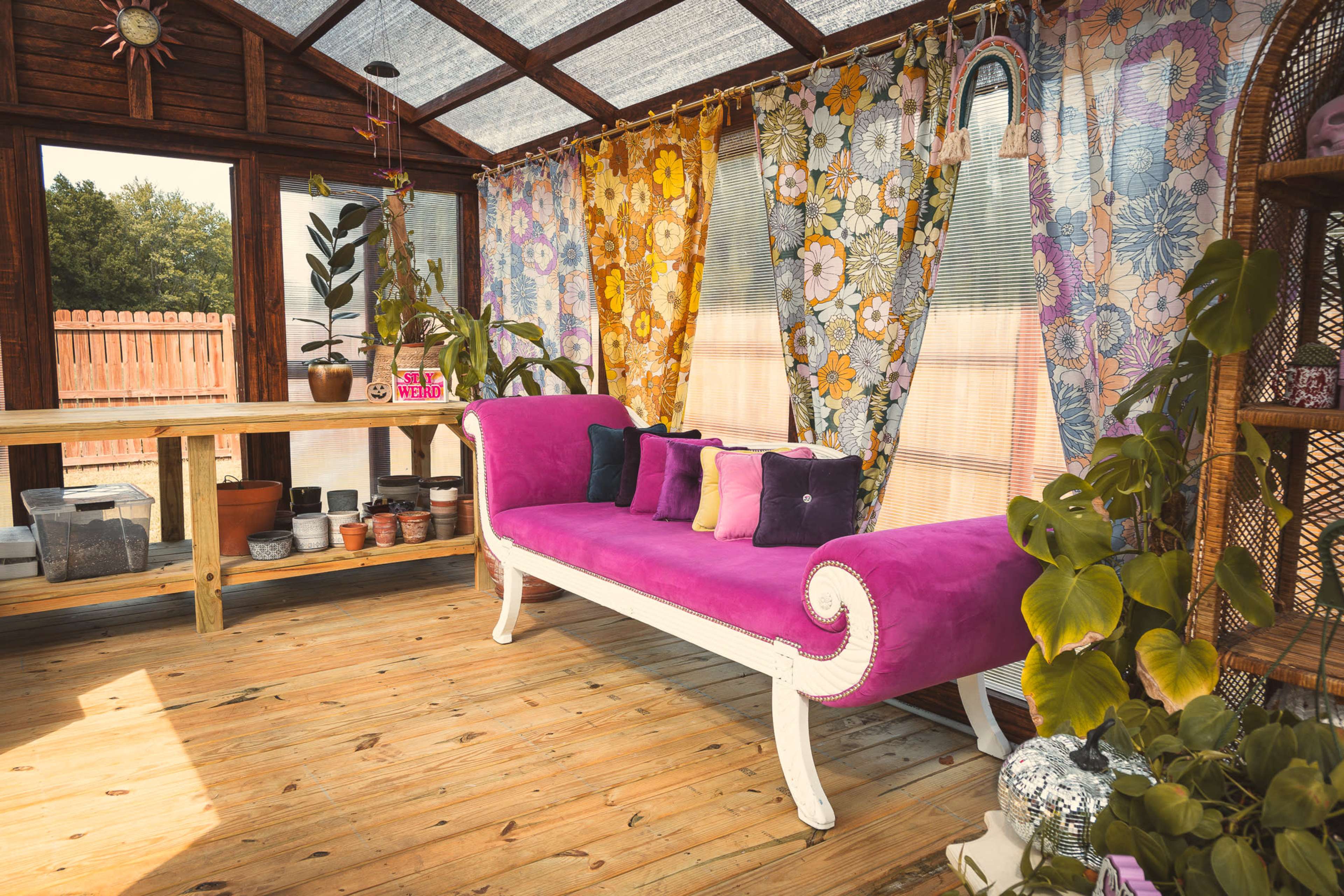 A colorful greenhouse features a pink sofa with decorative cushions and vibrant floral curtains, surrounded by potted plants on wooden shelves.