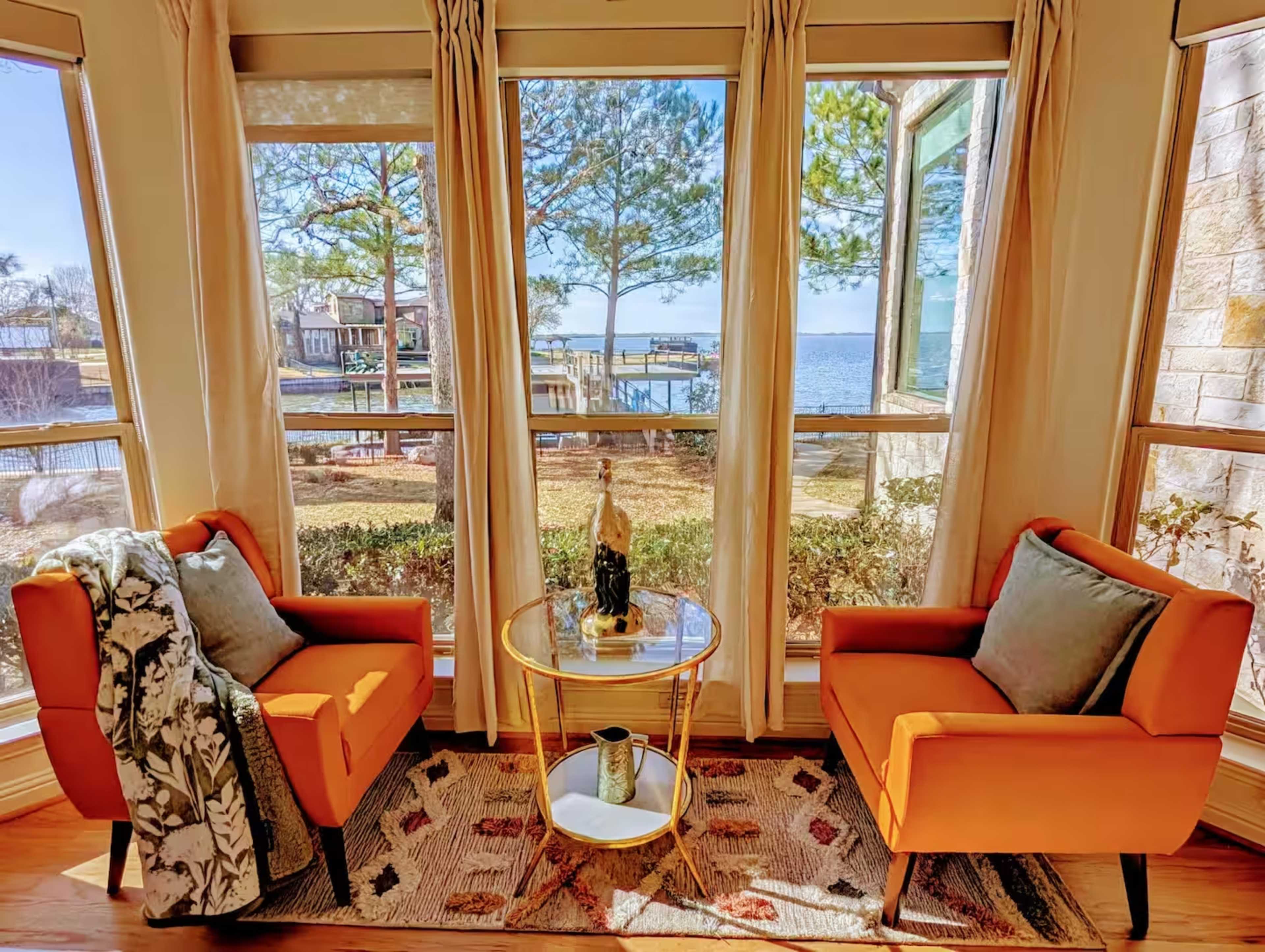 Two orange armchairs are positioned near large windows that overlook a lake, with a small round table between them and decorative items on the table.