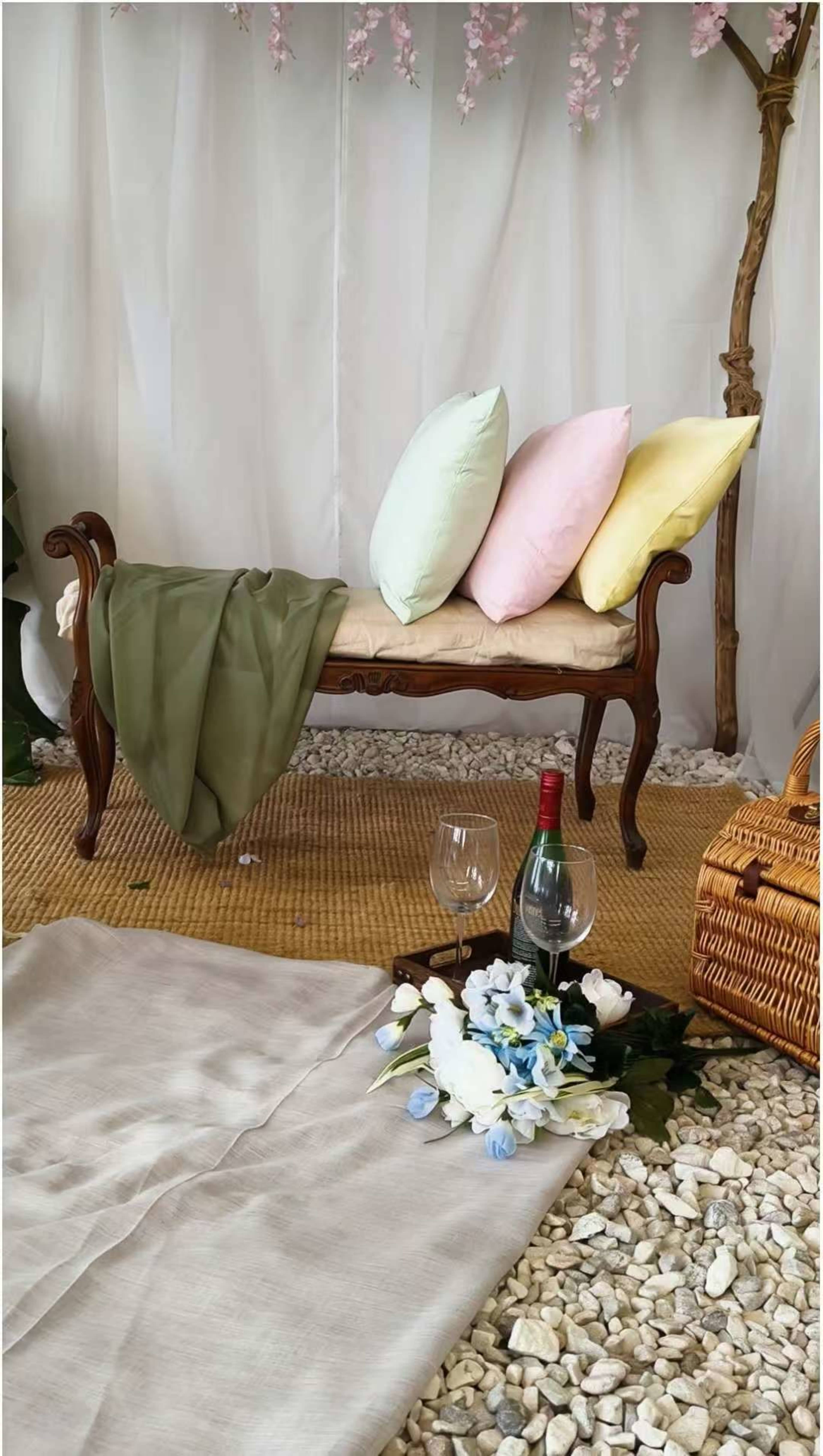 A wooden bench with four colorful pillows sits on a textured rug surrounded by decorative flowers and a bottle of wine with two glasses.