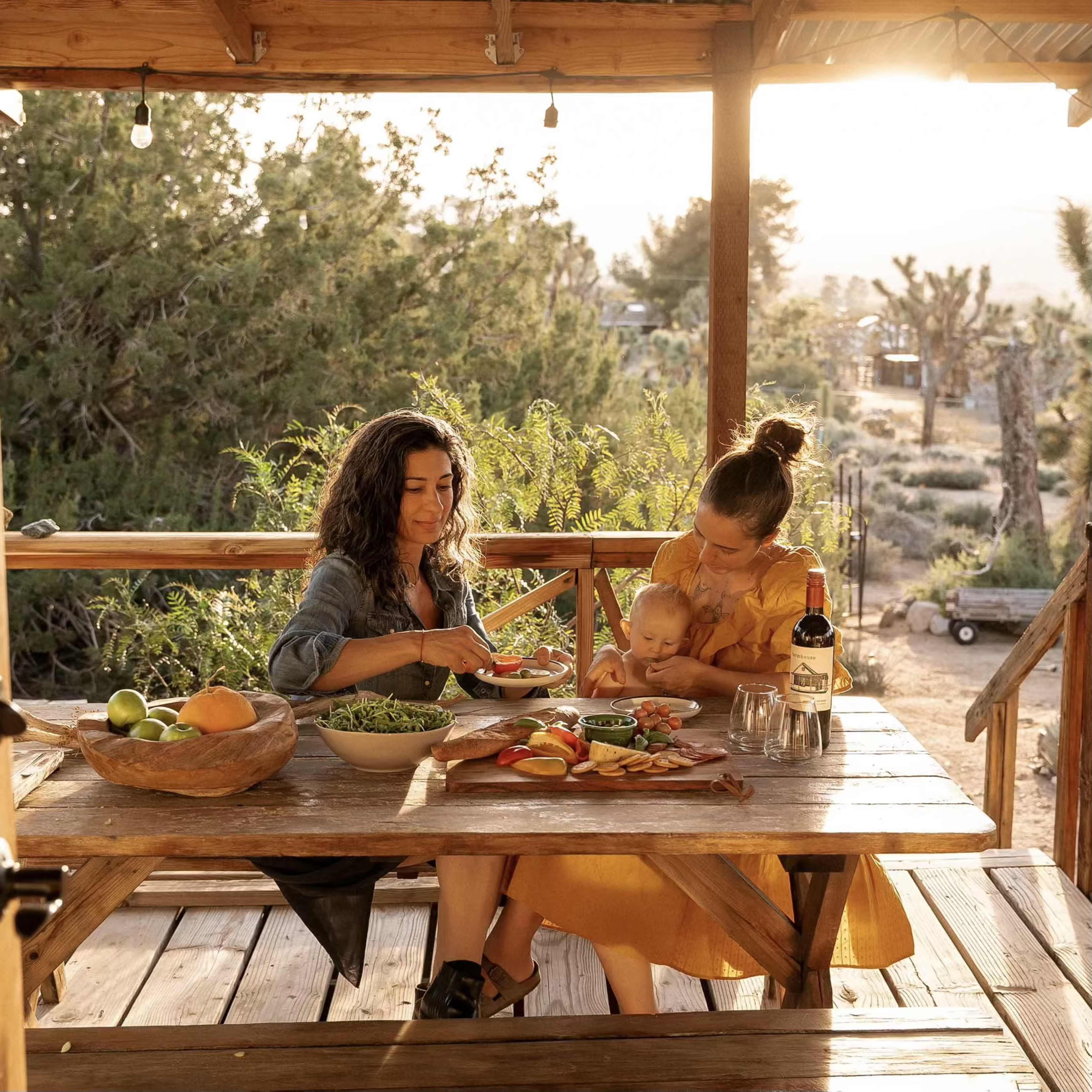 Two women and a baby are seated at a wooden table outdoors, enjoying a meal with a spread of fruits and dishes under a warm, golden light.