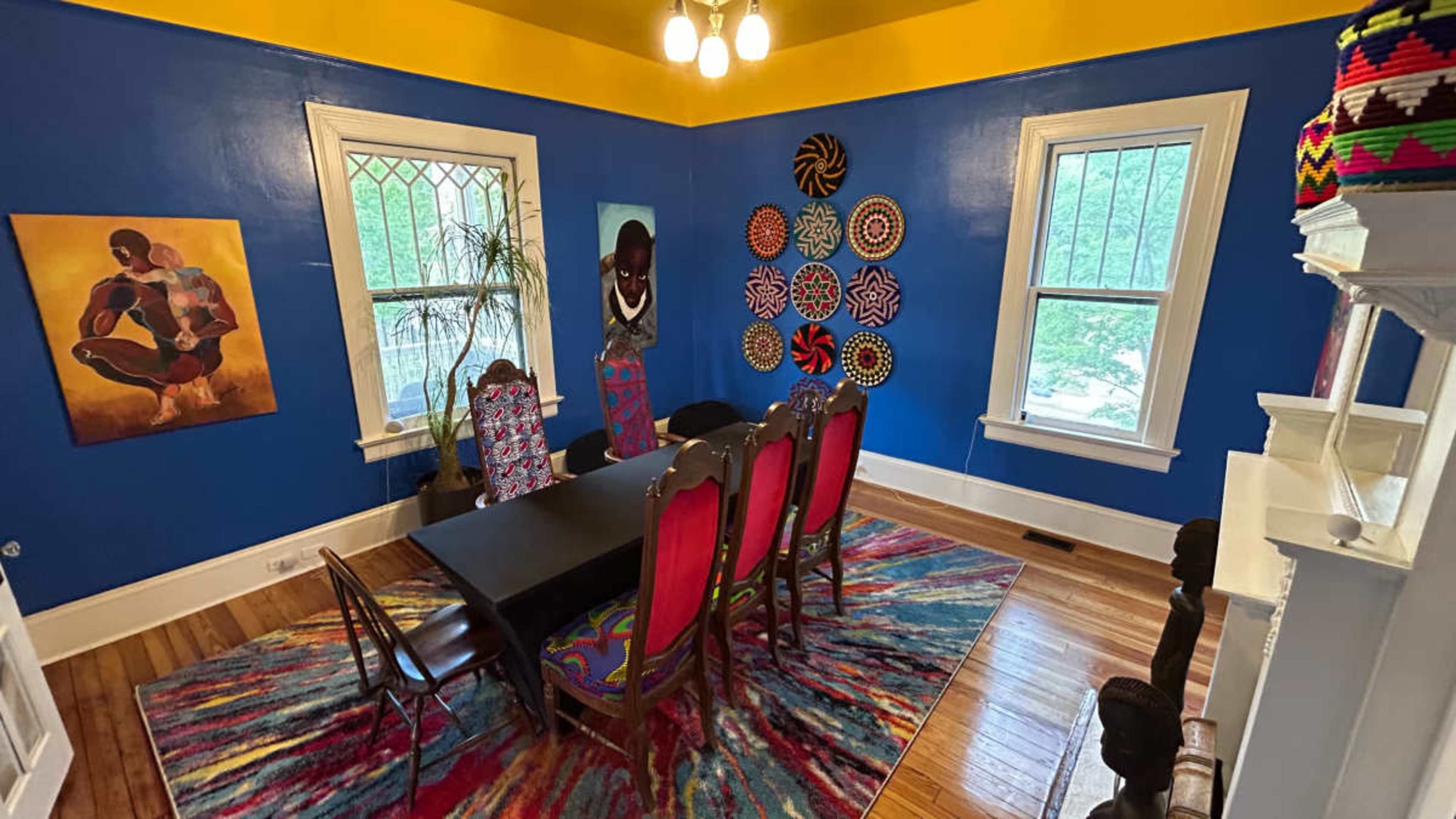 The image shows a colorful dining room featuring a black table surrounded by six chairs, with vibrant wall art and a patterned rug on the wooden floor.