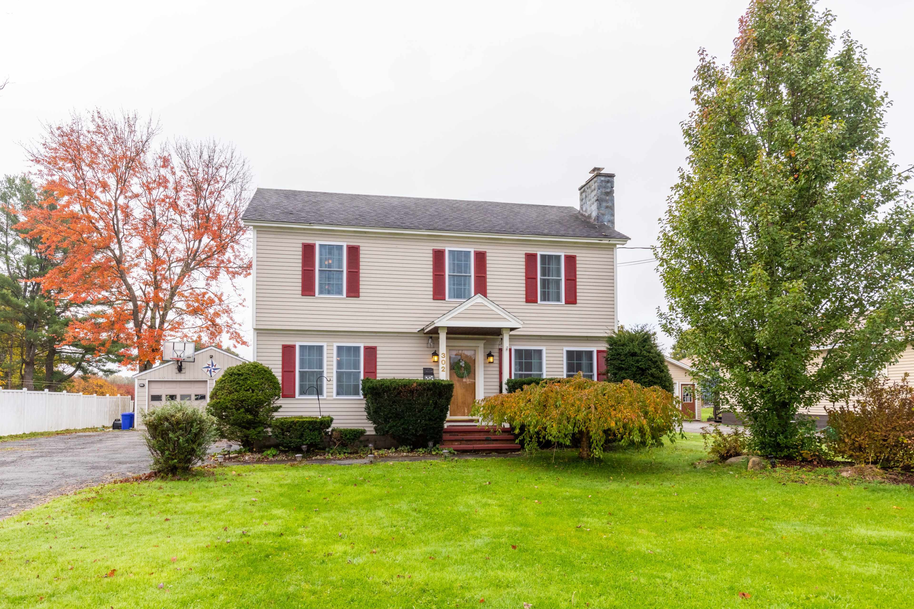 A two-story house with red shutters sits on a green lawn, surrounded by trees and a gravel driveway.