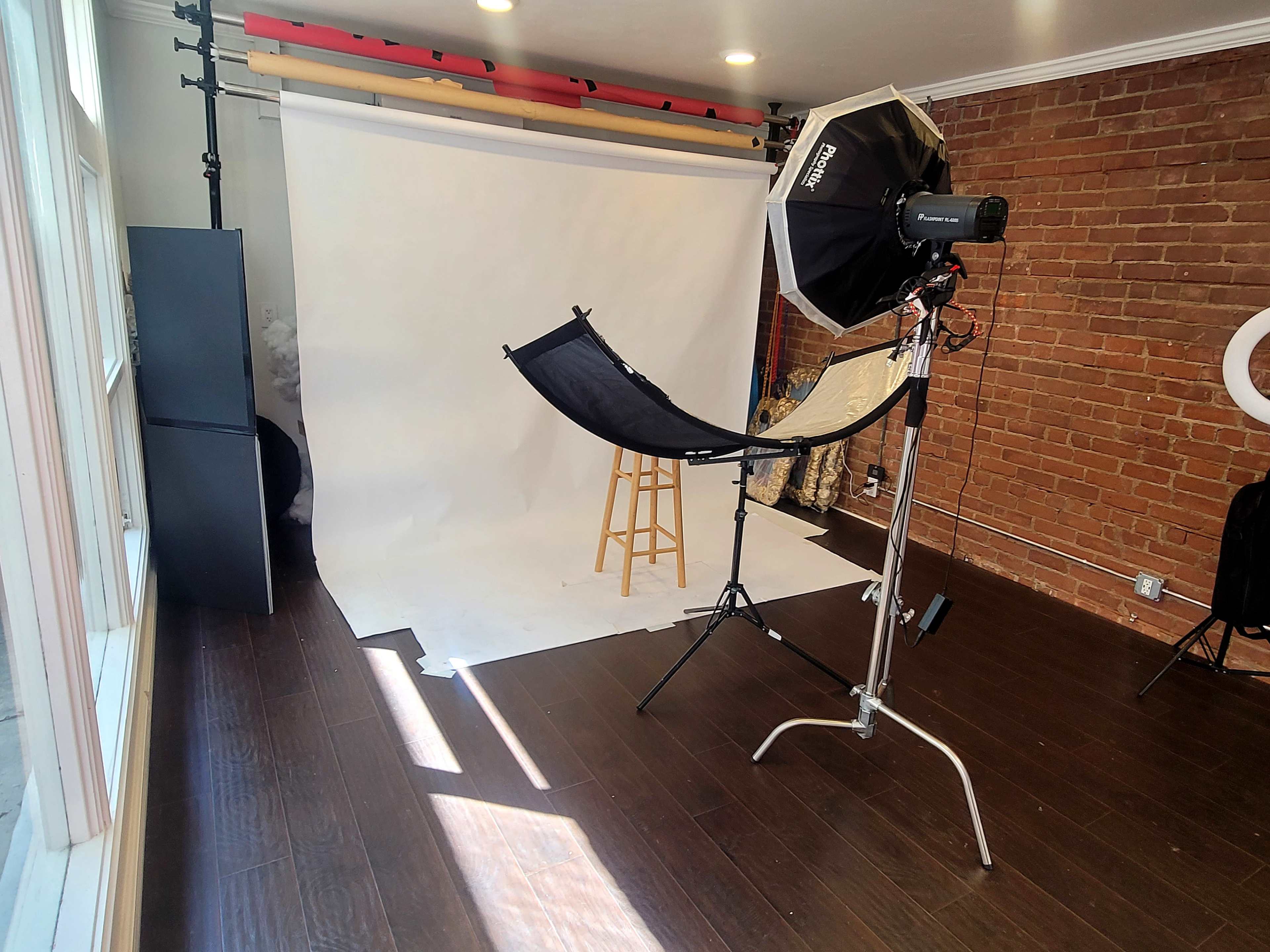 The image shows a photography studio setup featuring a white backdrop, a stool, lighting equipment, and a dark wood floor.