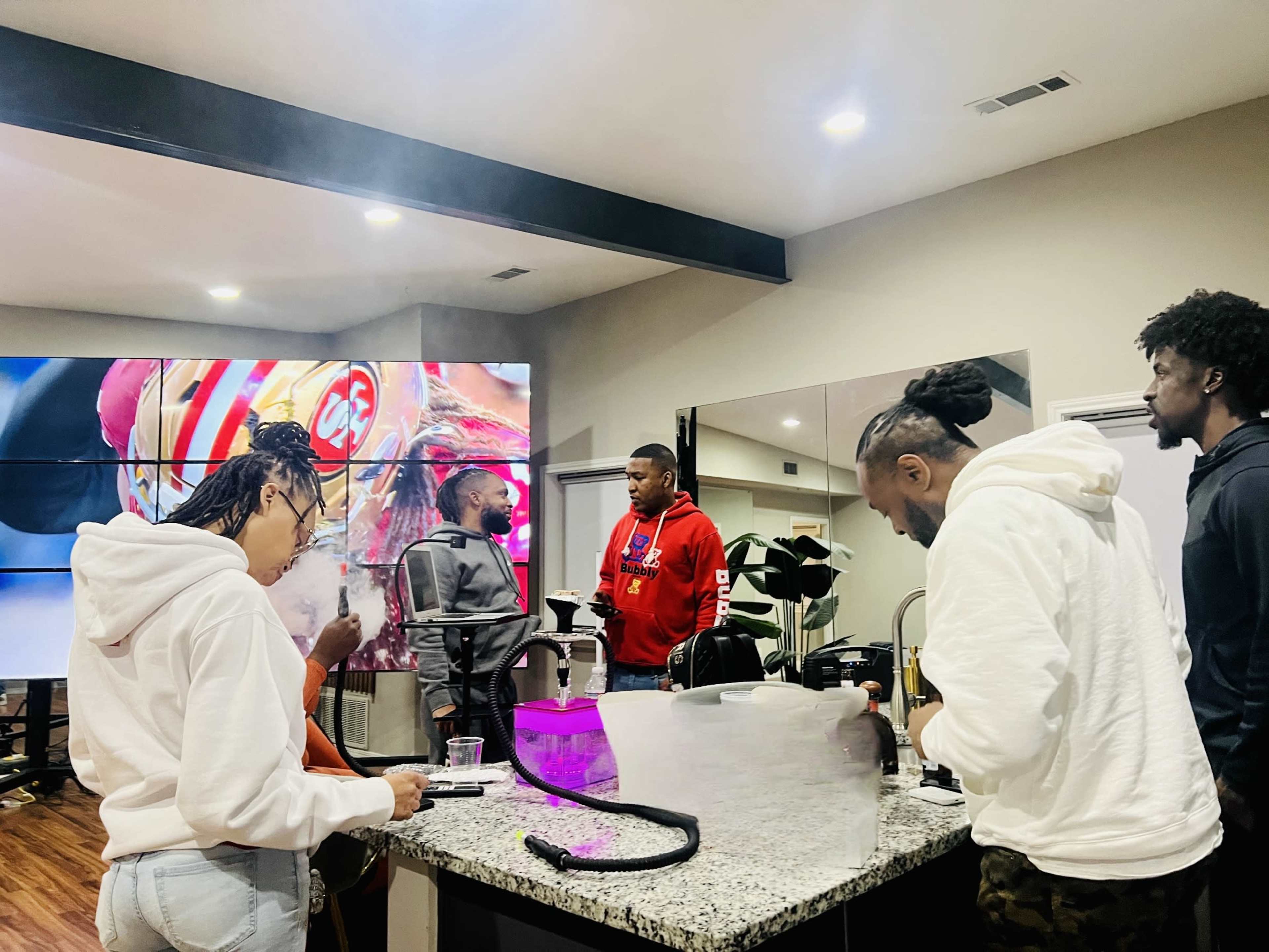A group of four people is gathered around a kitchen island in a modern interior, with a large screen displaying images in the background.