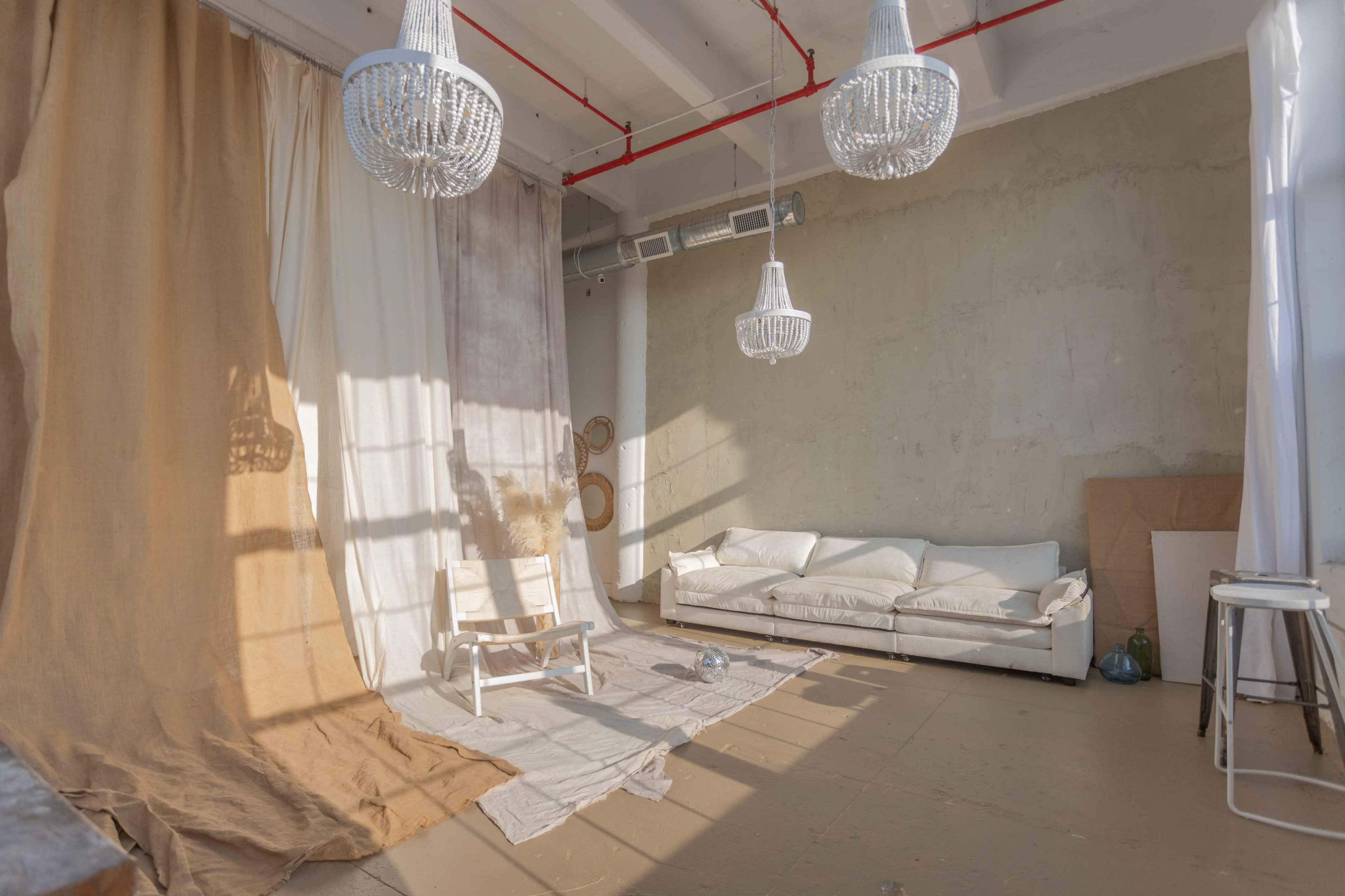 A bright, minimalistic room with a white sofa, a light-colored floor covering, and three hanging chandeliers, illuminated by natural light.