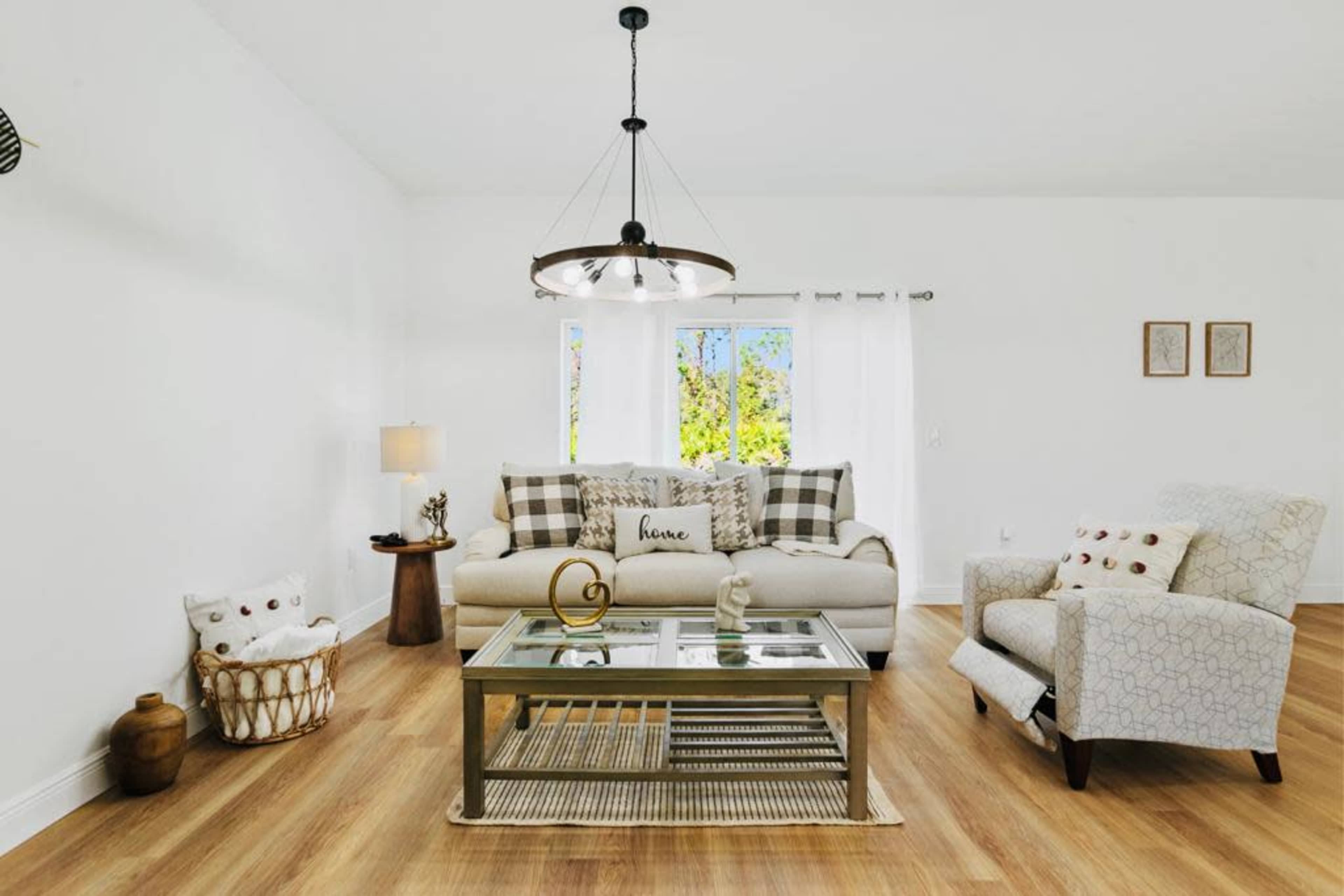 The image shows a bright, modern living room with a sofa, an armchair, a glass coffee table, and large windows allowing natural light to enter.