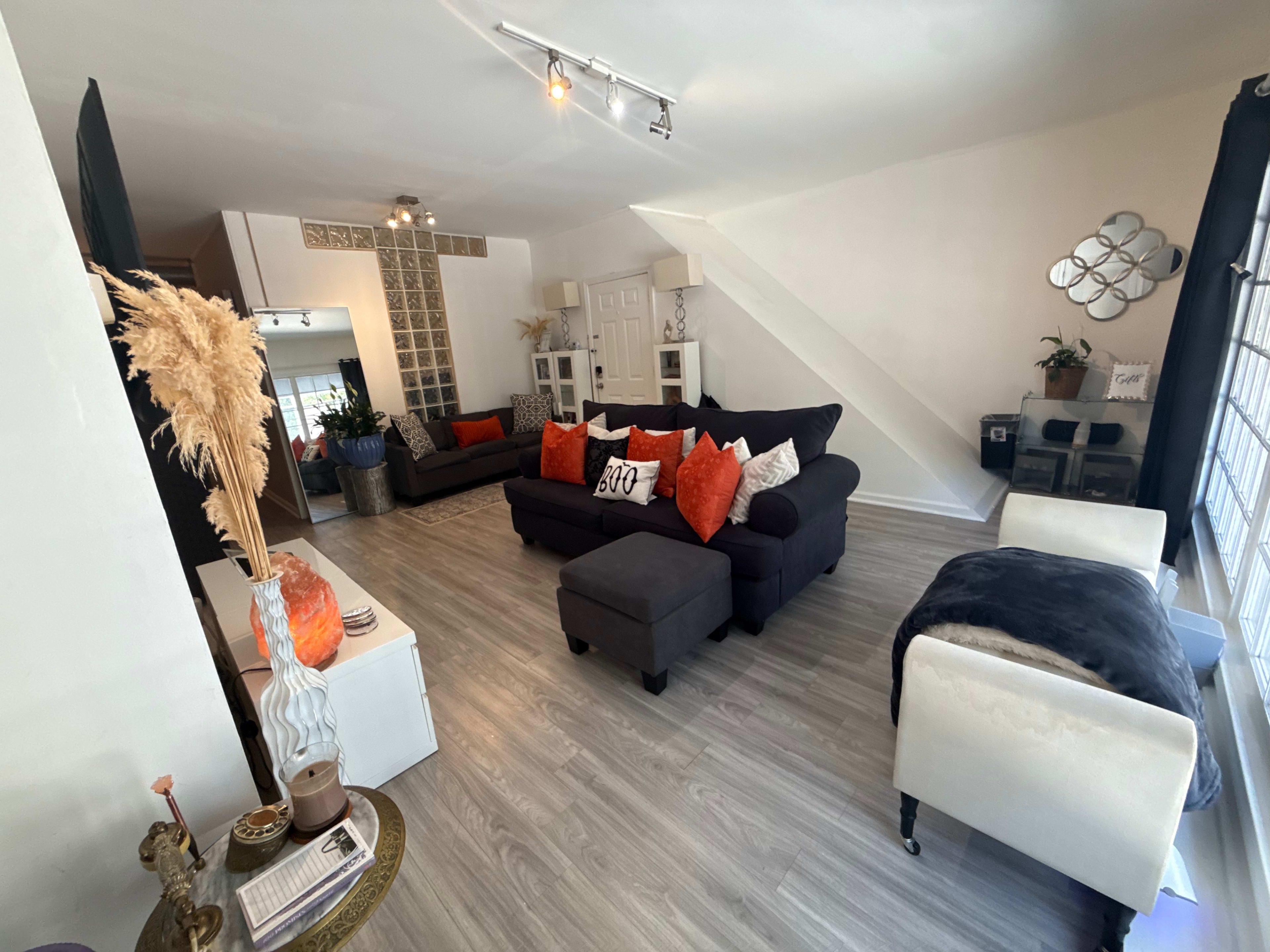 The image shows a spacious living room featuring a mix of dark and light furniture, with orange decorative pillows and an area rug contrasting the light wood flooring.