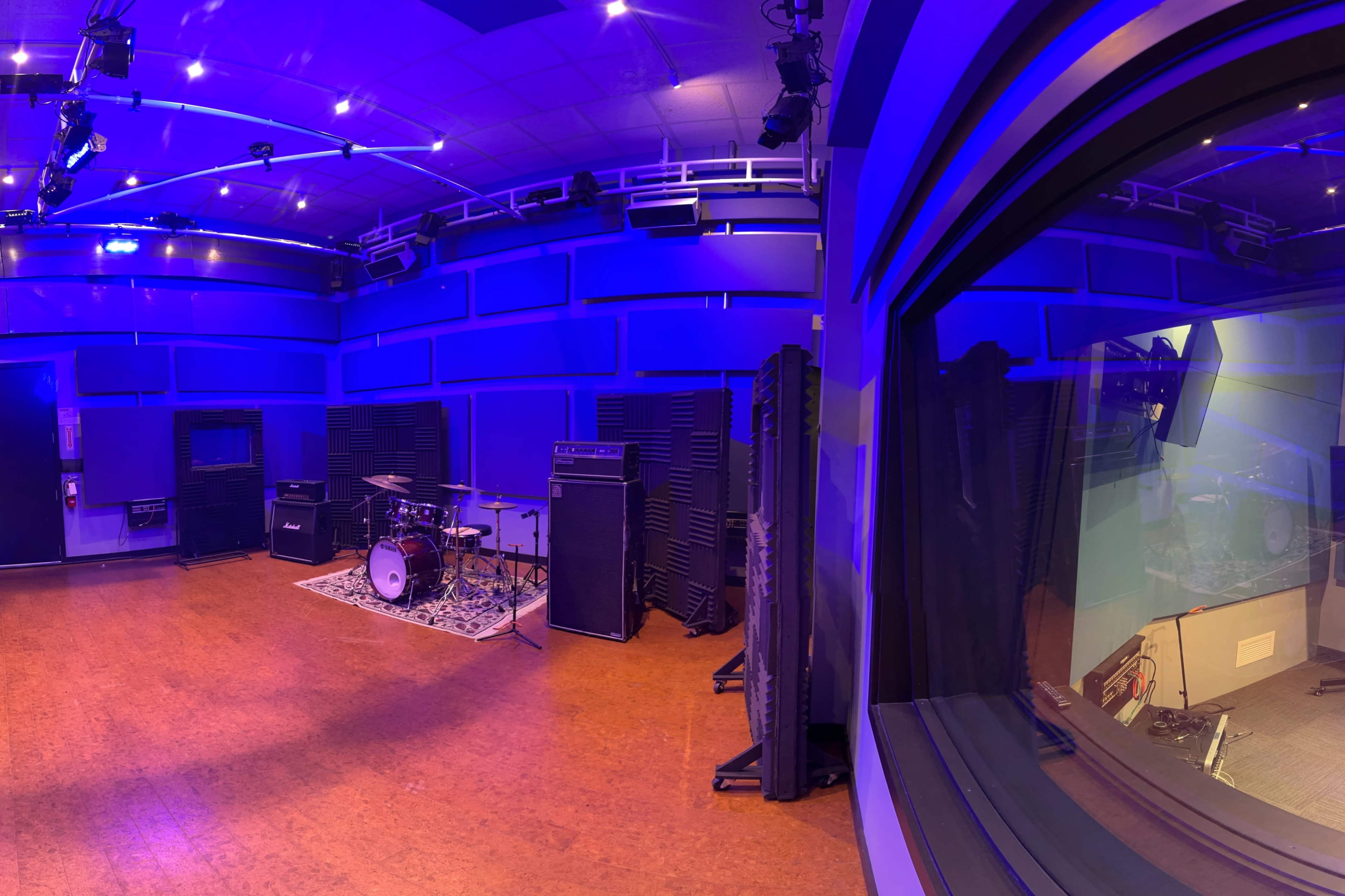 The image shows a sound studio with a drum kit and amplifiers in a blue-lit room, viewed through a glass panel from an adjacent control room.