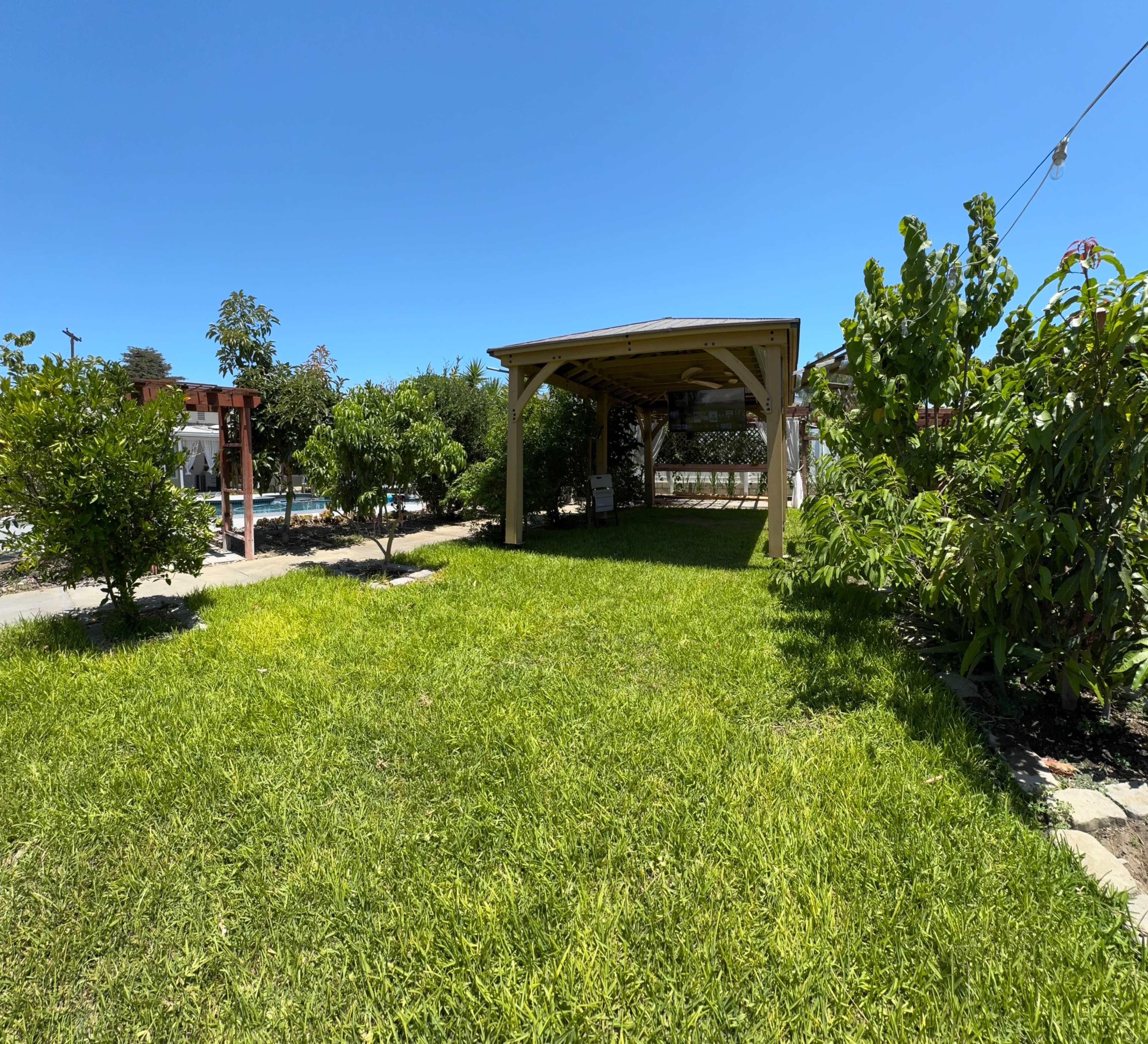 A grassy area with a wooden gazebo and surrounding trees under a clear blue sky.