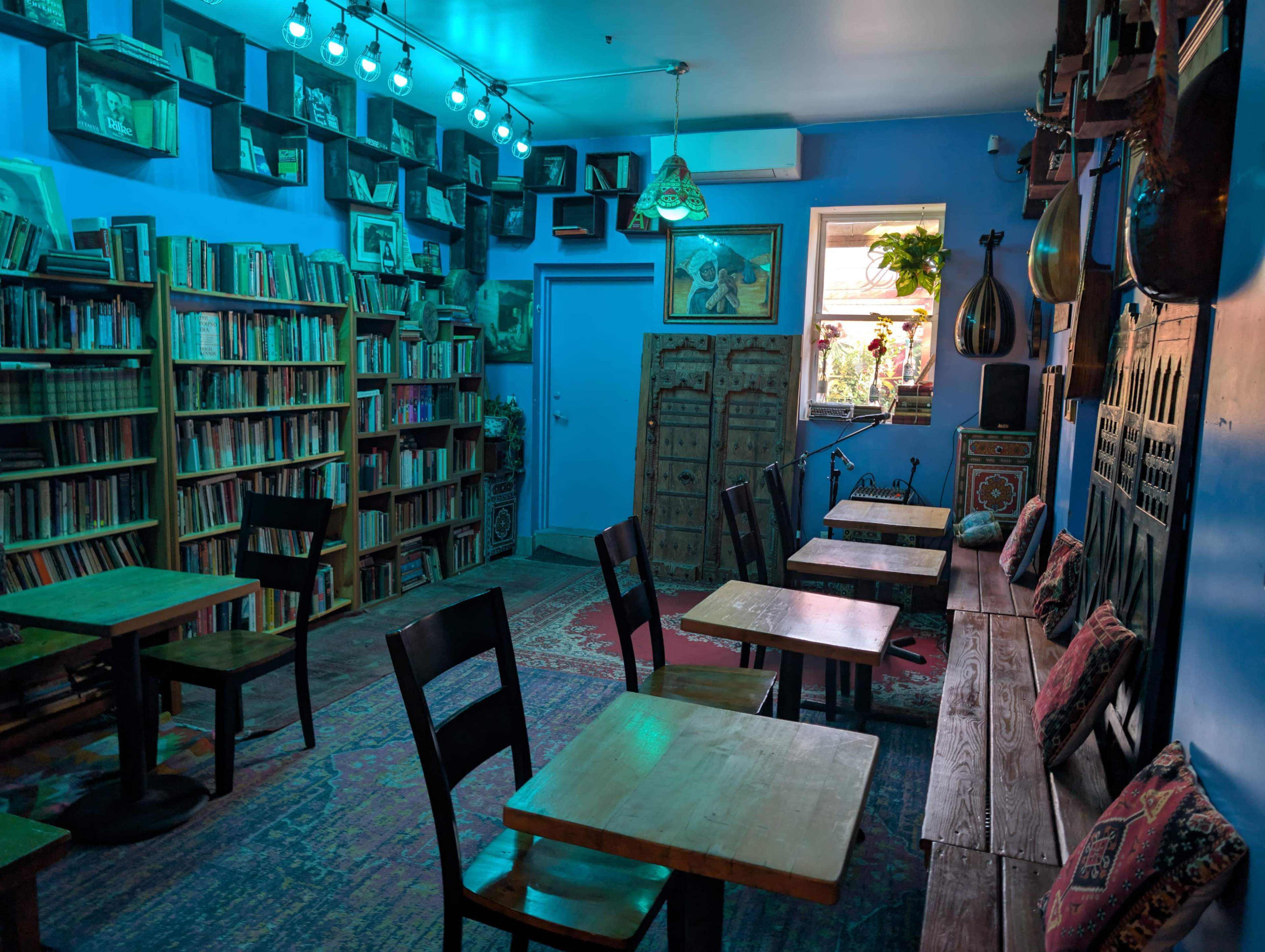 The image shows a cozy, book-filled café with wooden tables and chairs, set against walls painted in blue and adorned with shelves and decorative items.