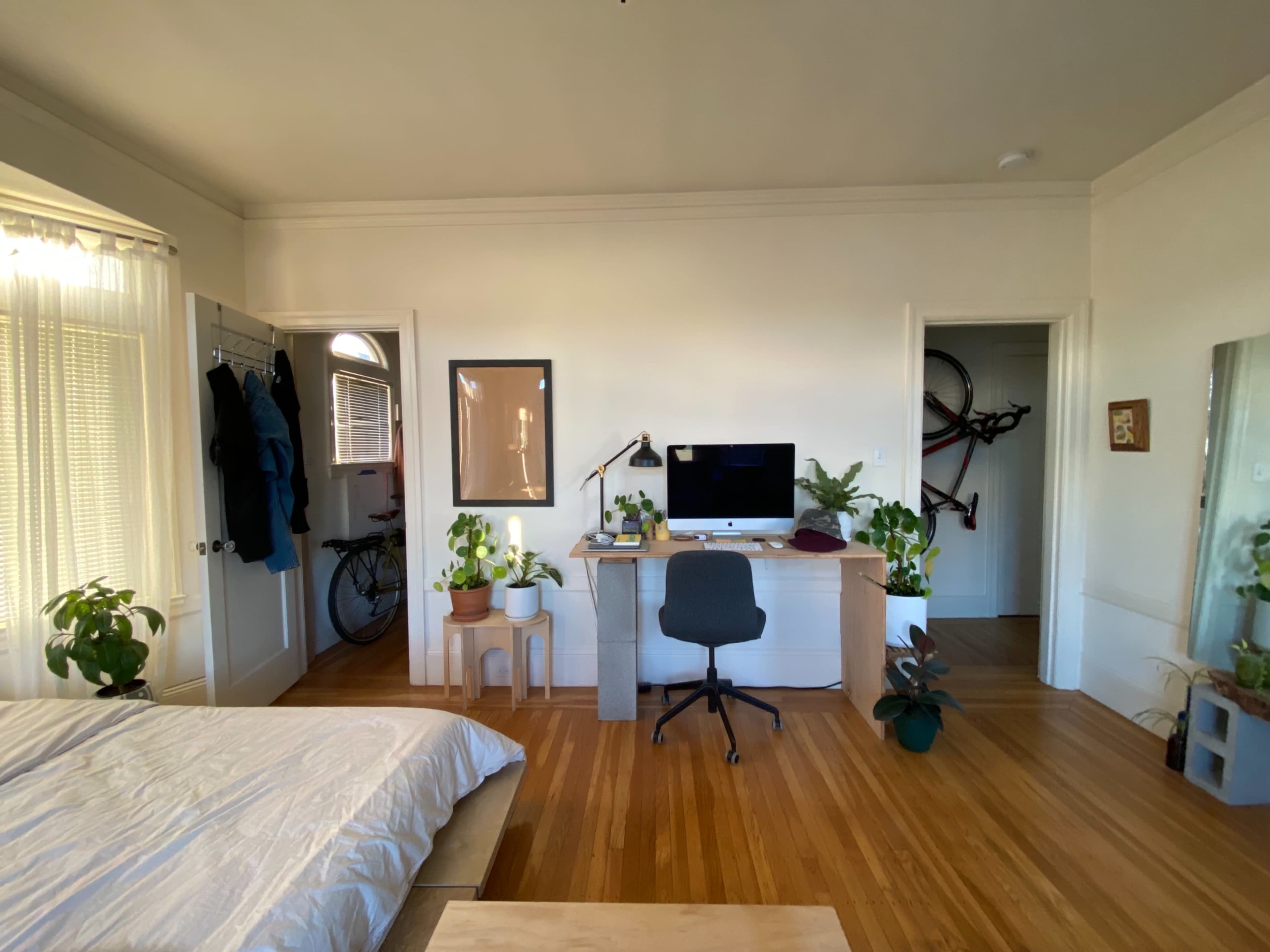 A bedroom features a bed, a desk with a computer, several potted plants, and a bicycle hanging on the wall.