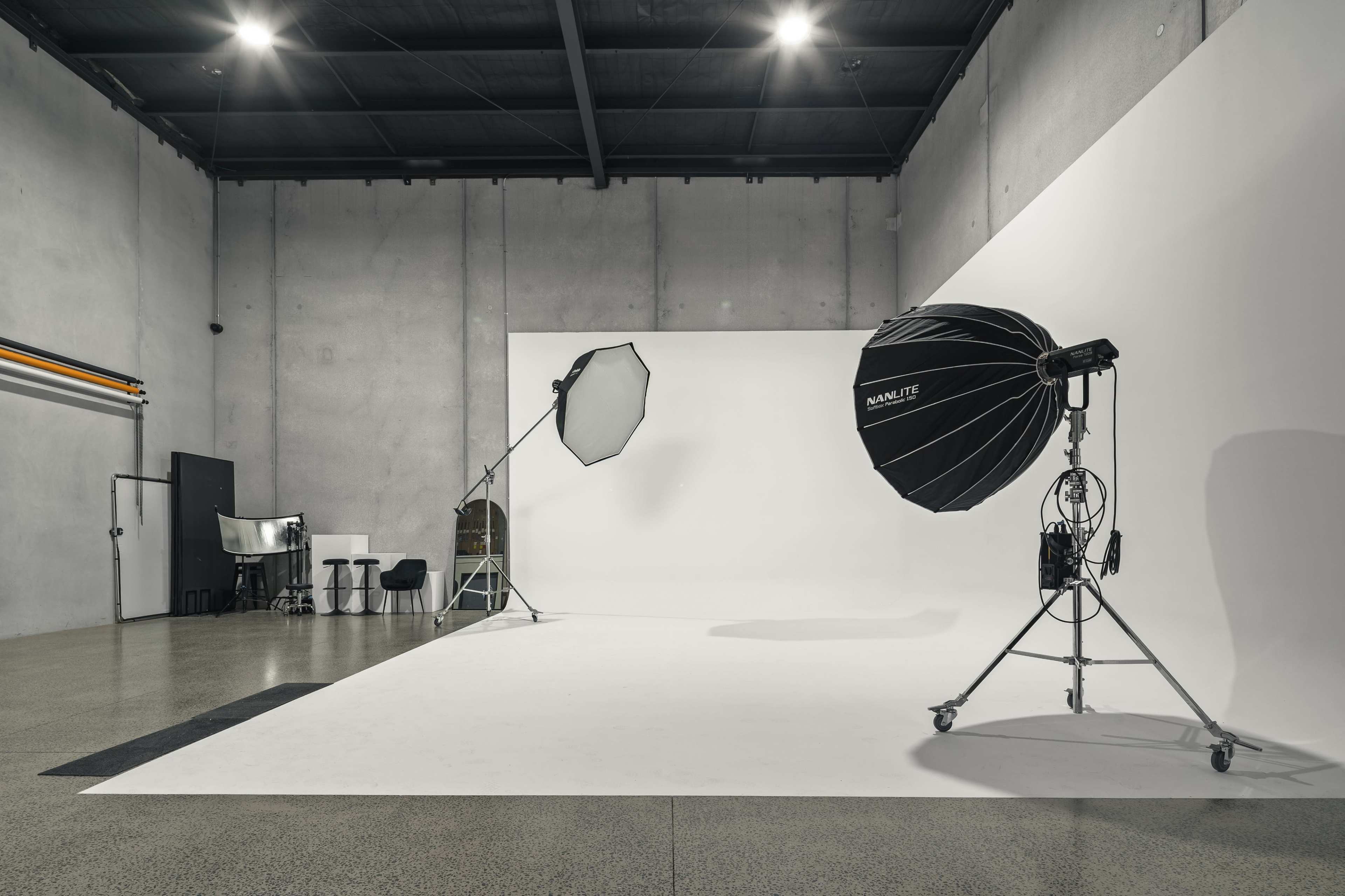 The image shows a photography studio featuring a large white backdrop, various lighting equipment, and a few chairs and tables.