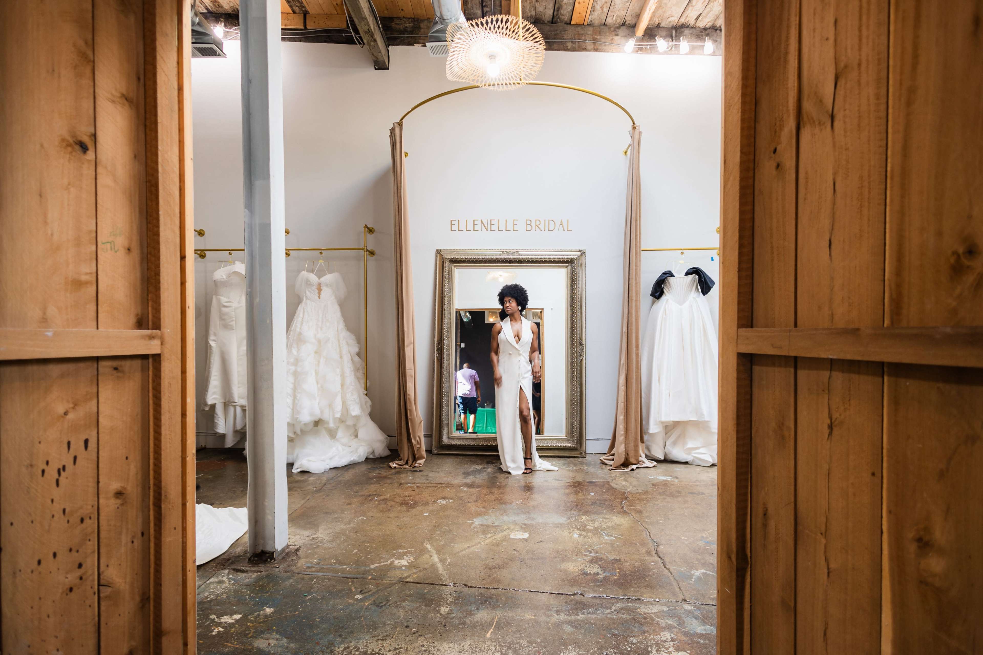 A woman stands in front of a large mirror in a bridal shop, surrounded by various wedding dresses displayed on racks.