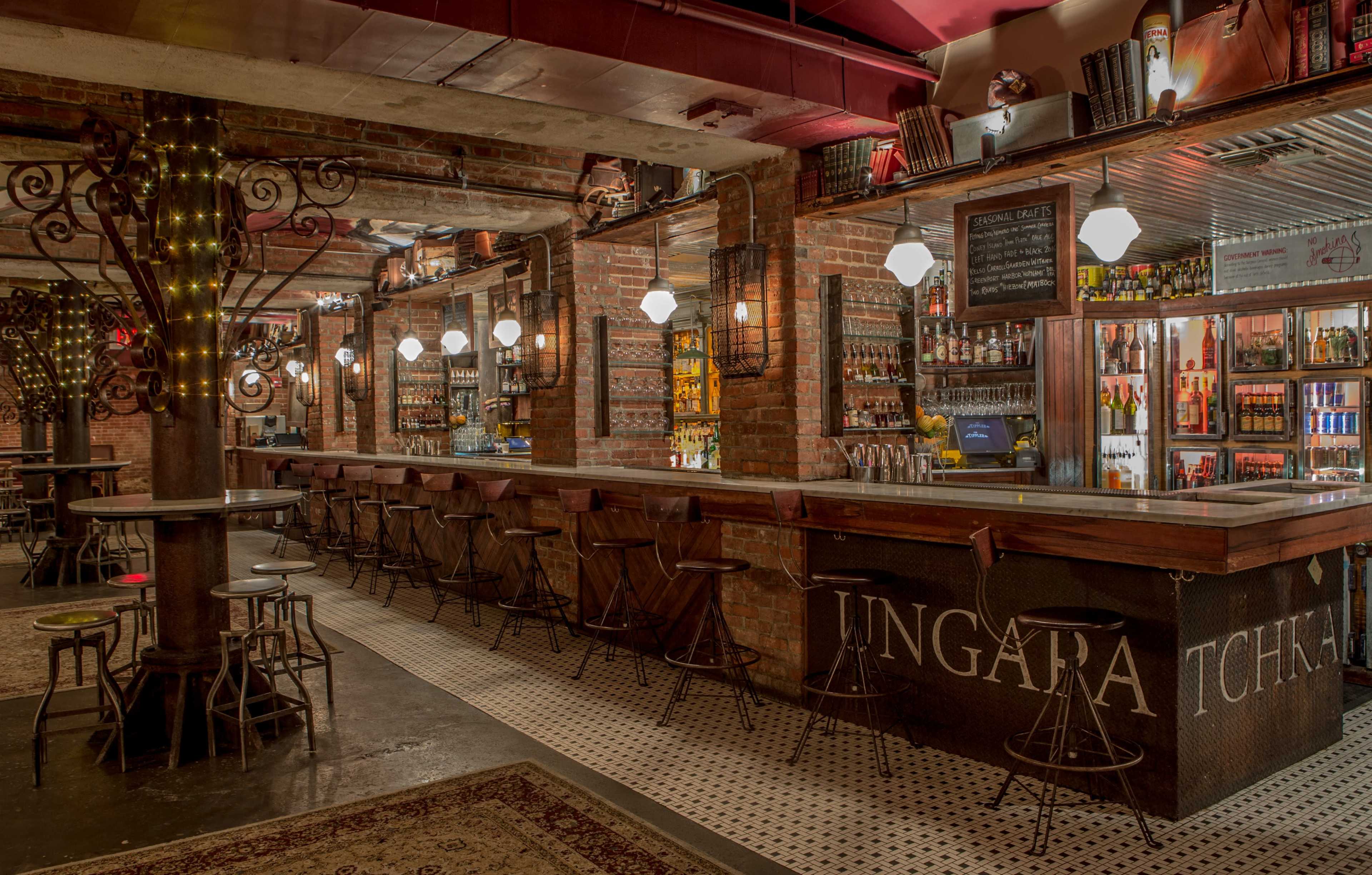 The image shows a dimly lit bar with a wooden counter, metal stools, and exposed brick walls decorated with shelves of bottles.