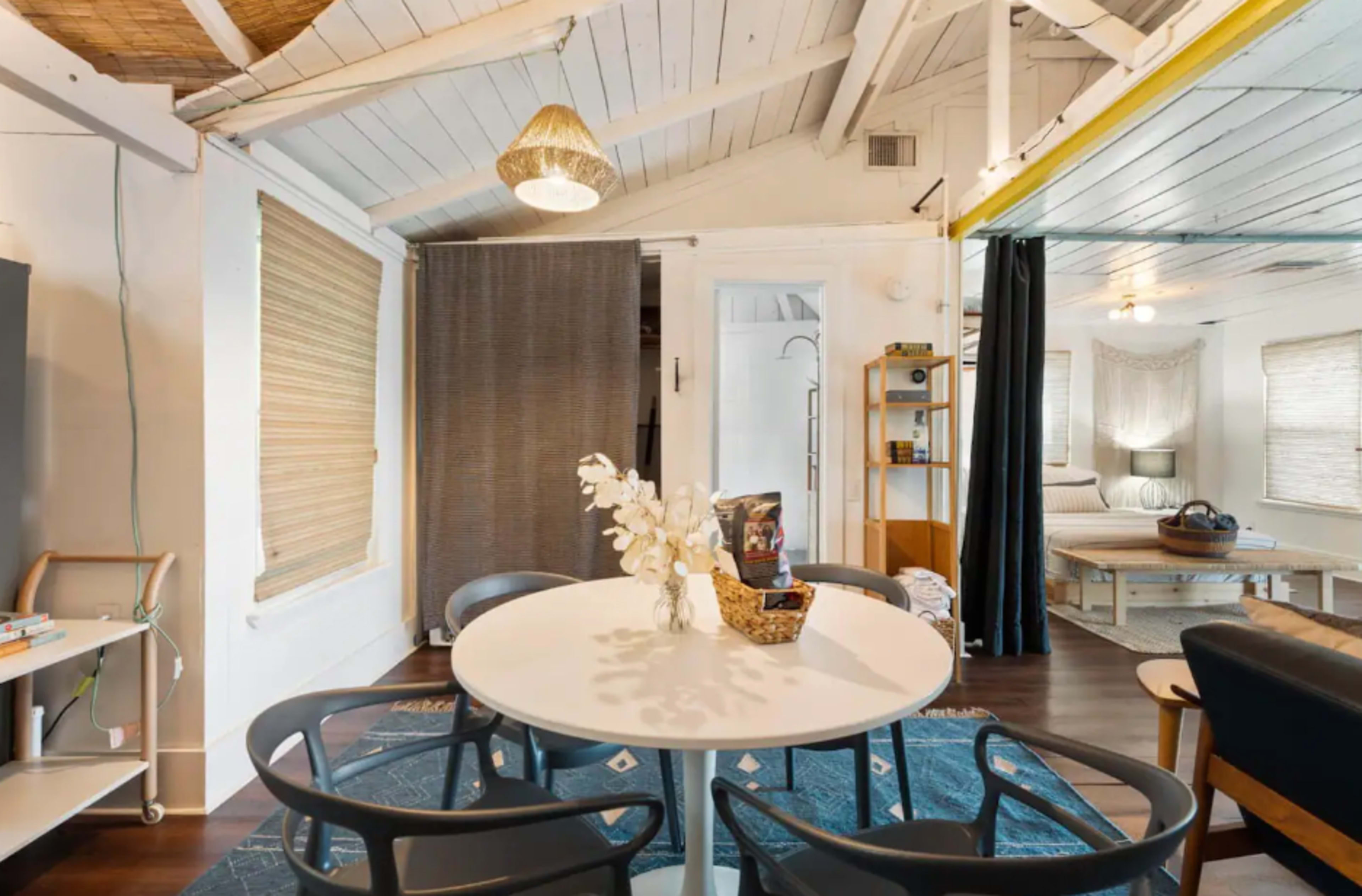 A bright, open living space with a round dining table surrounded by black chairs, and separated areas featuring a kitchen and a seating area.