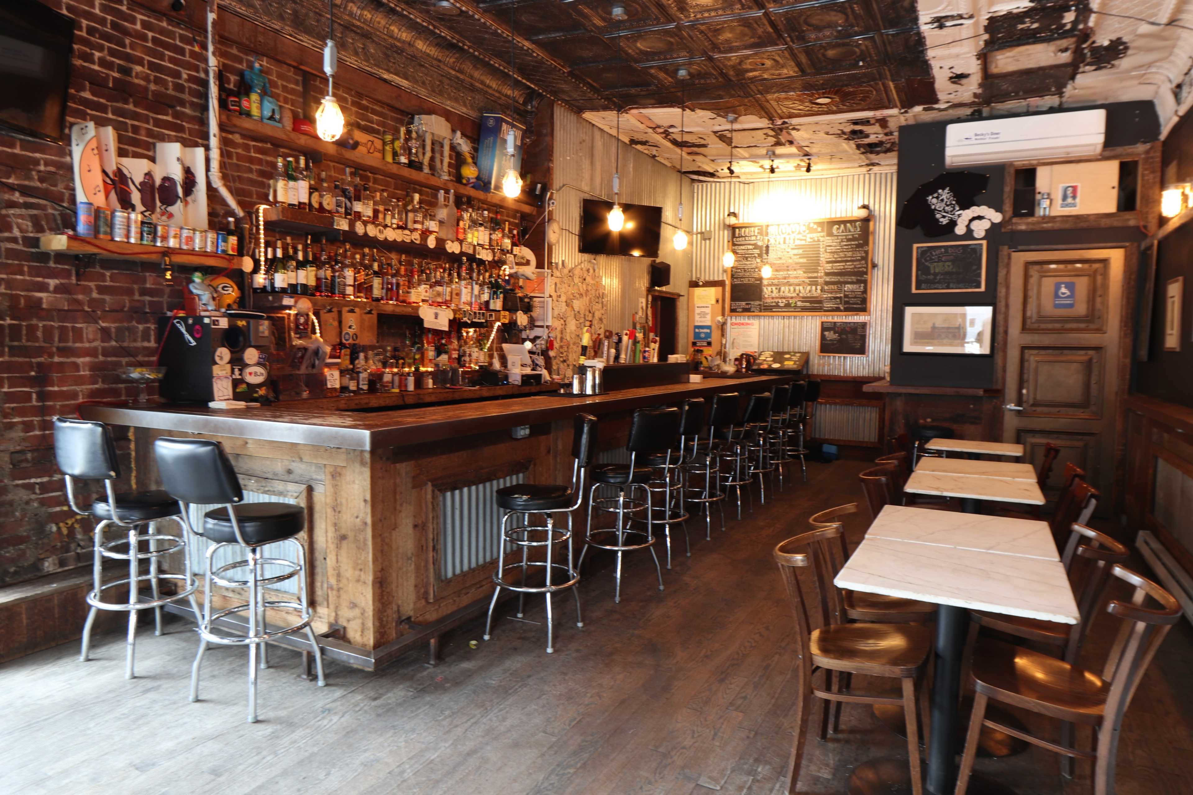 A rustic bar interior with a long wooden counter, several bar stools, and shelves stocked with various bottles.