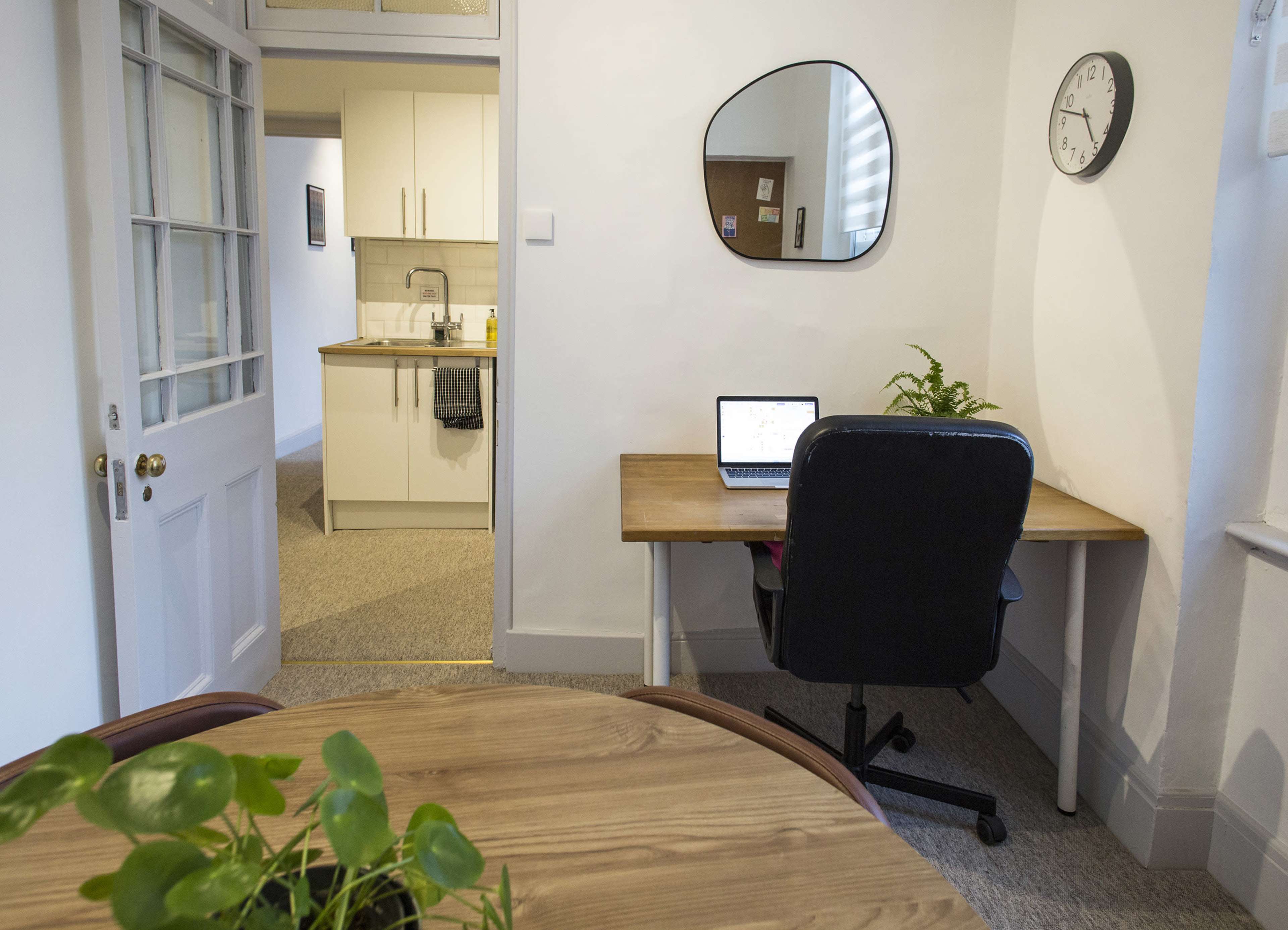 A small home office setup featuring a desk with a laptop, a chair, and a mirror, with a kitchen area visible in the background.