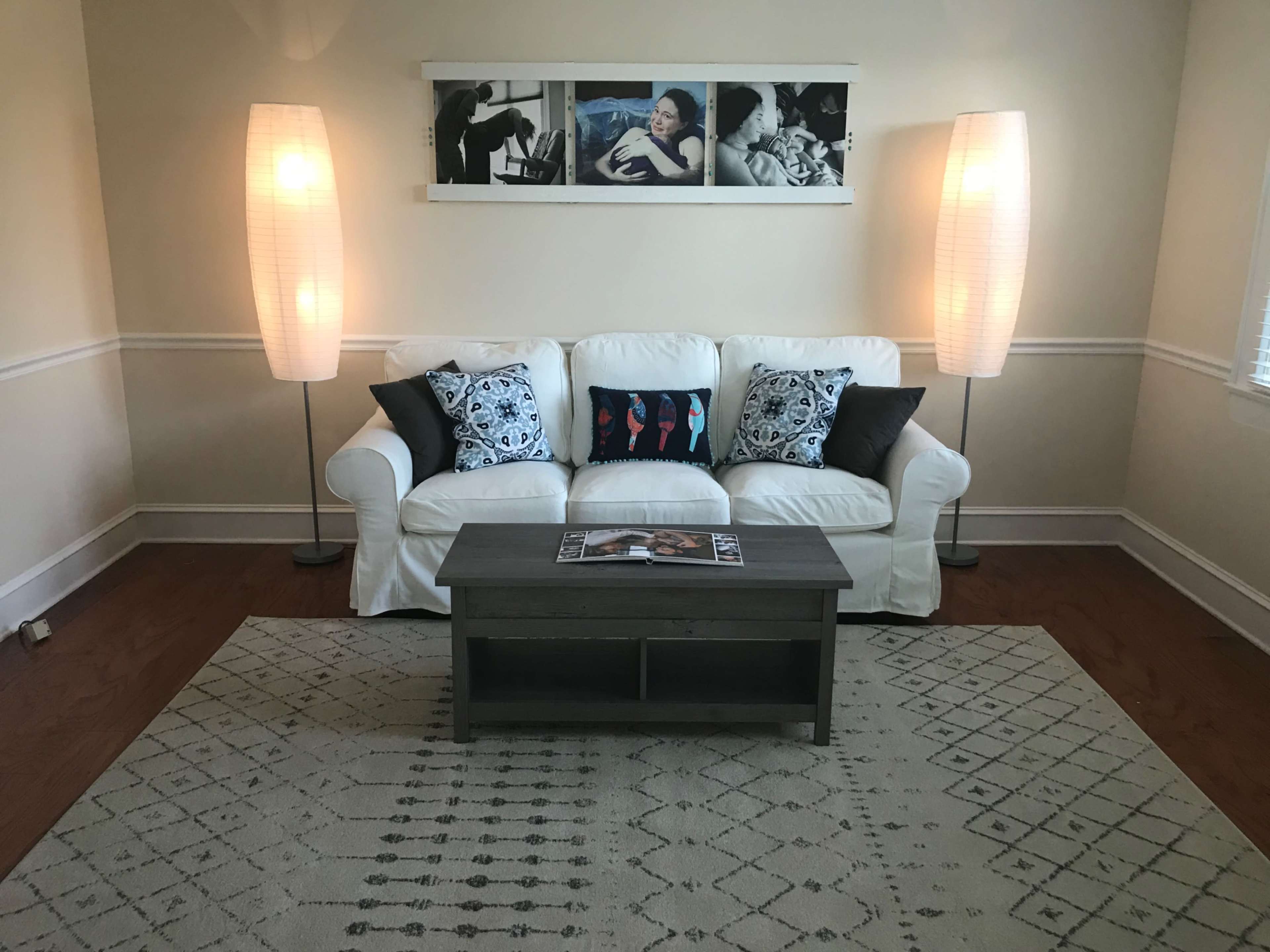 A white couch with decorative pillows sits in a living room, flanked by two floor lamps, with a coffee table in front and a framed image on the wall behind.