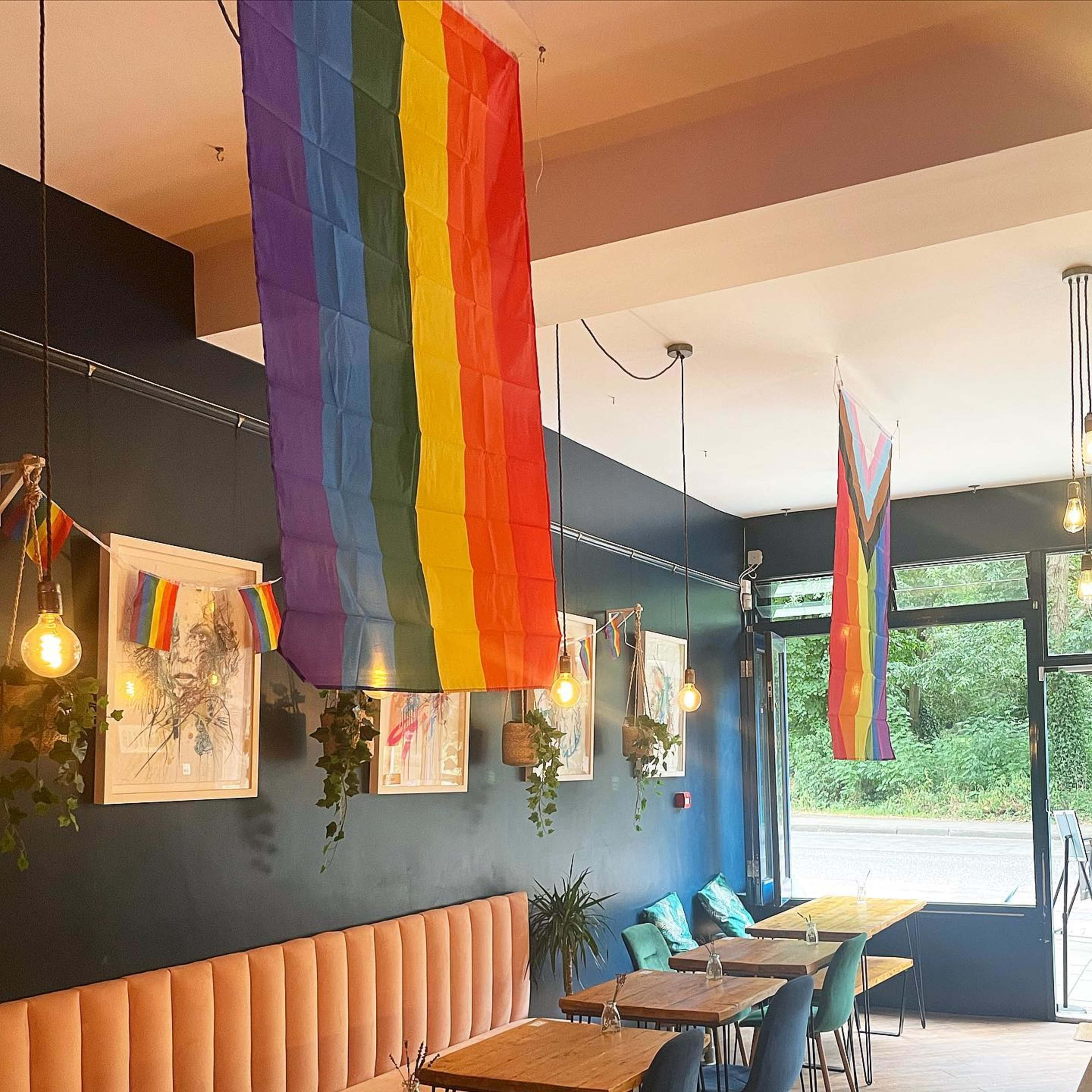 The image shows a brightly lit café interior decorated with rainbow flags and artwork, featuring tables and seating along the walls.