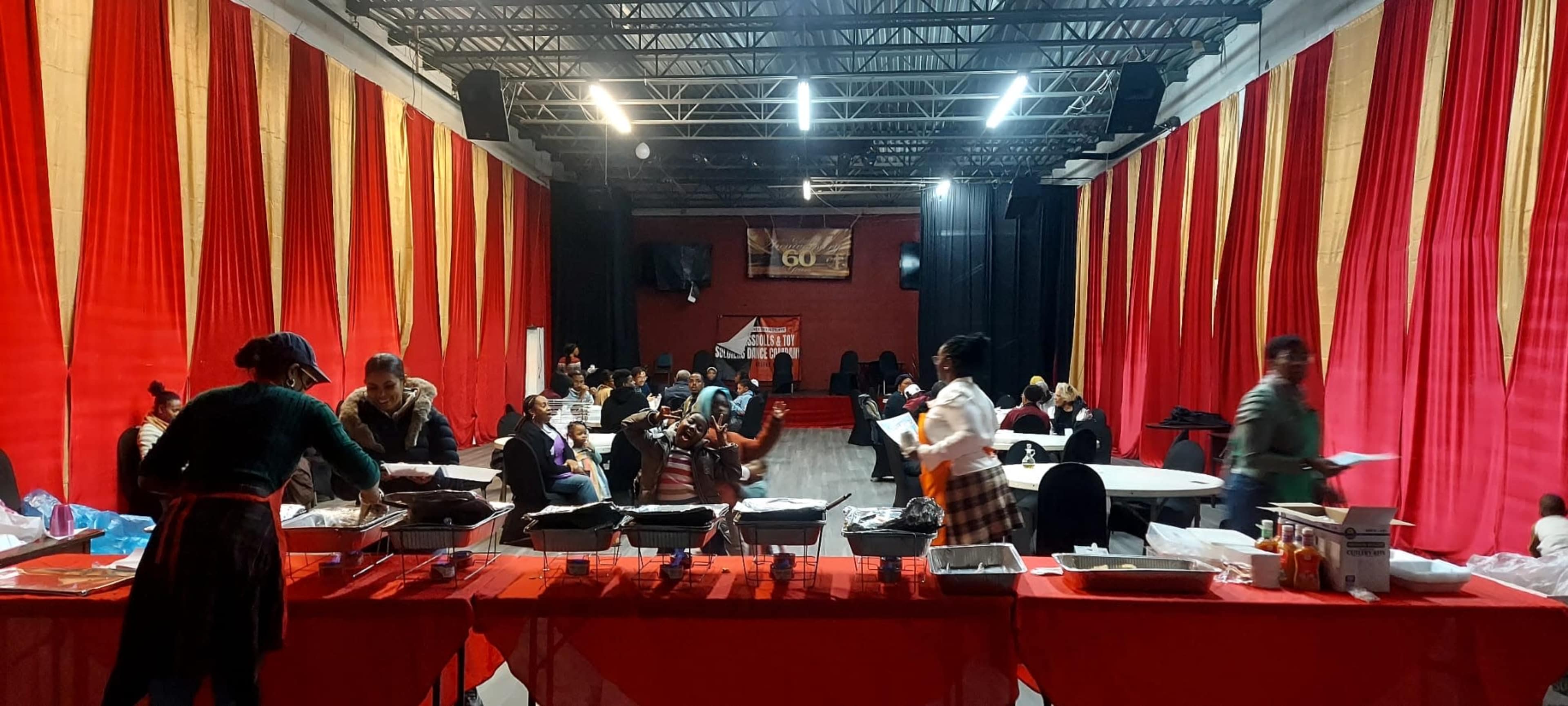 The image shows a large event space with red and gold draped curtains, where people are seated at tables and others are interacting or preparing food.