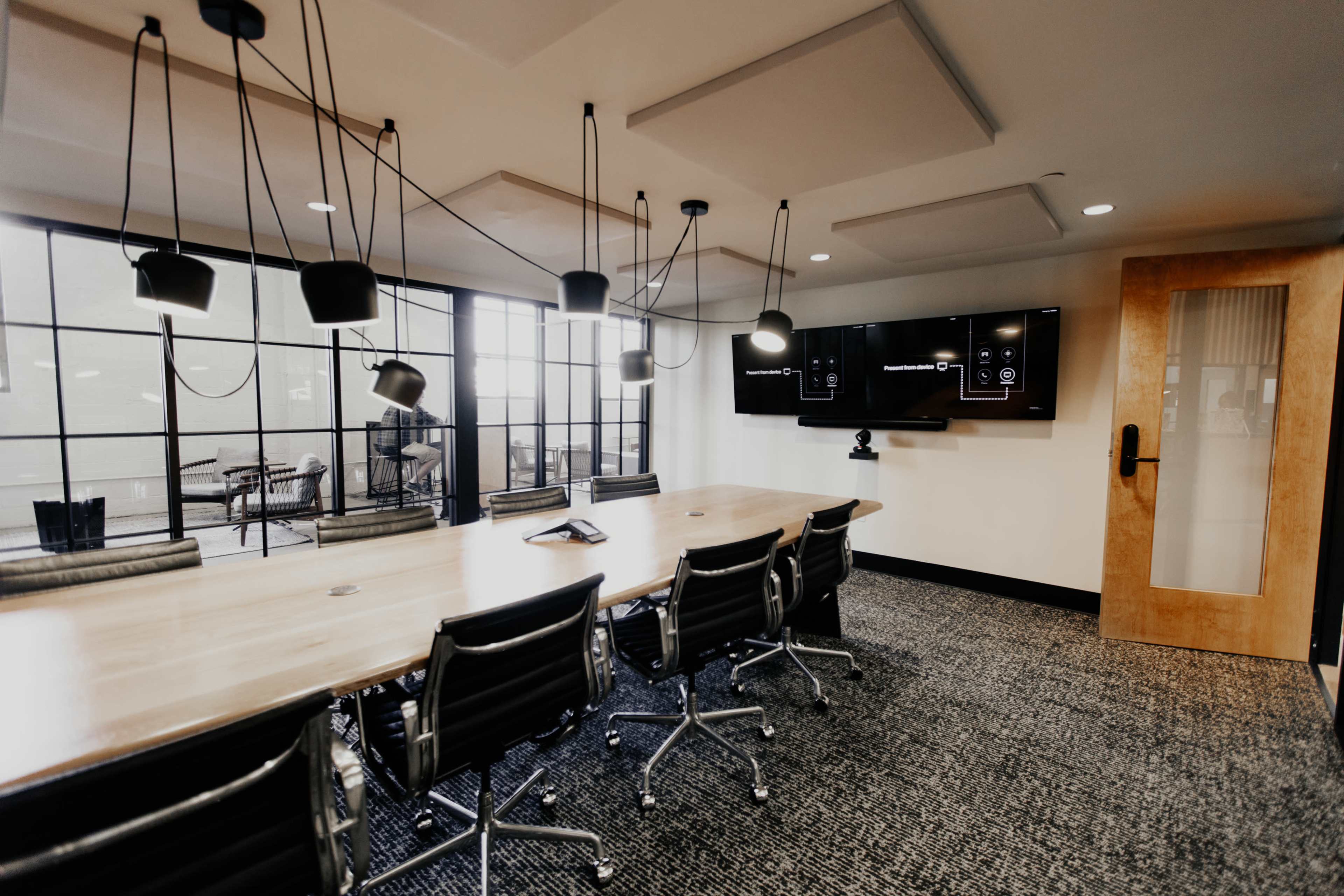 A modern conference room features a long wooden table surrounded by ergonomic chairs, with large windows, pendant lighting, and a wall-mounted screen.