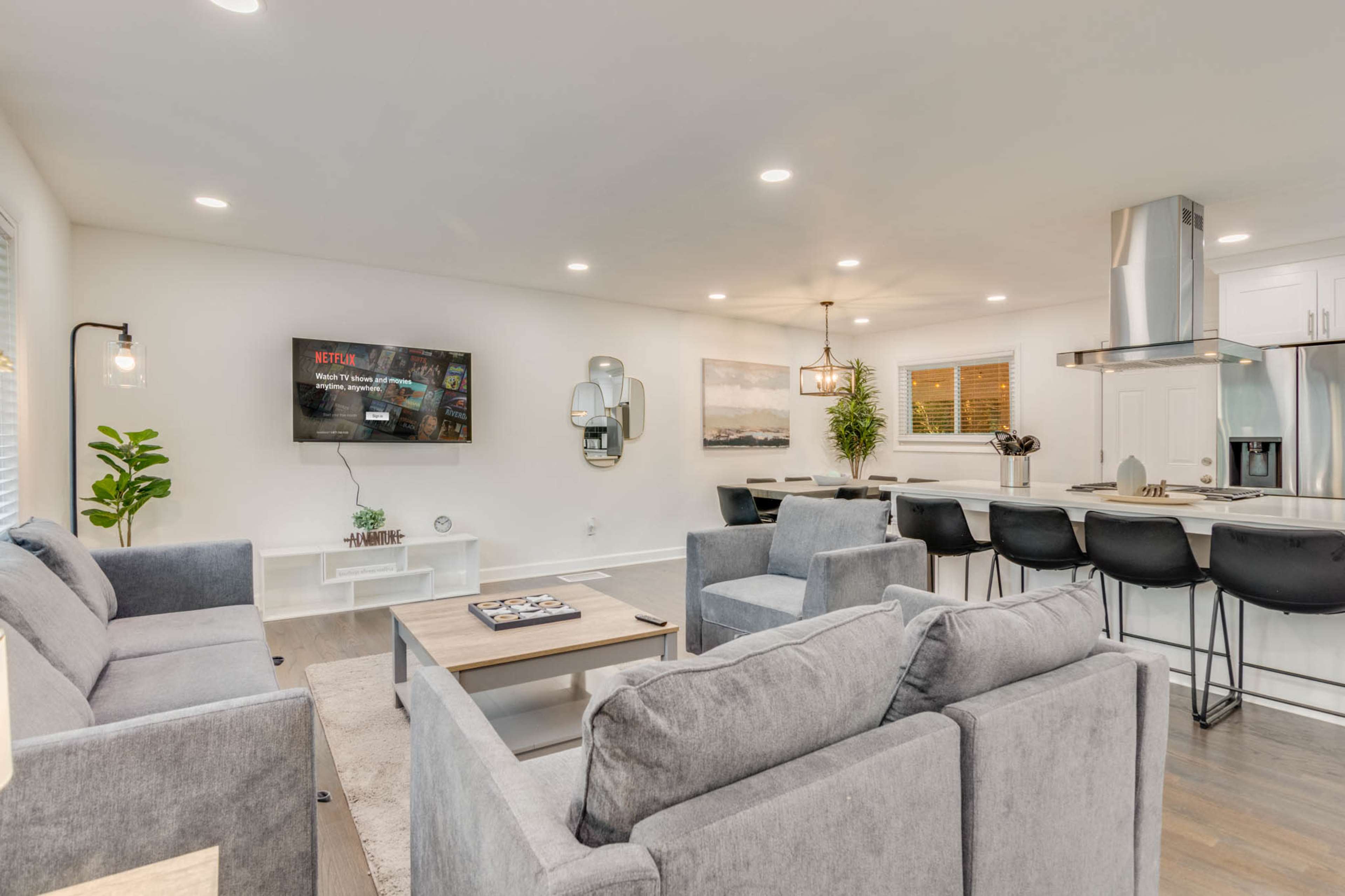 The image shows a modern, well-lit living space featuring a gray couch set, a wooden coffee table, and a kitchen area in the background with bar stools and a TV mounted on the wall.