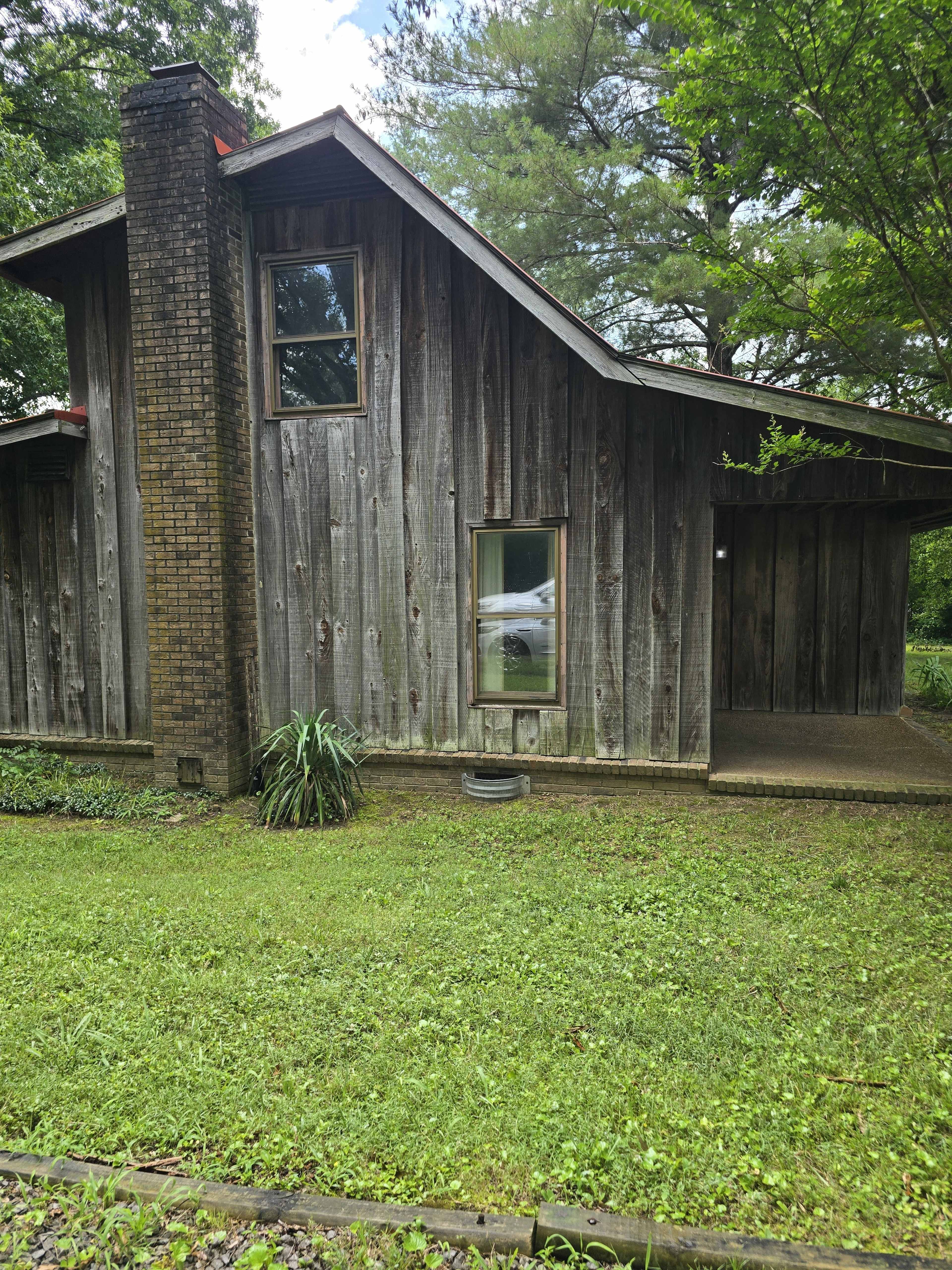 The image shows a weathered wooden house with a slanted roof and a small front porch, surrounded by grass and trees.