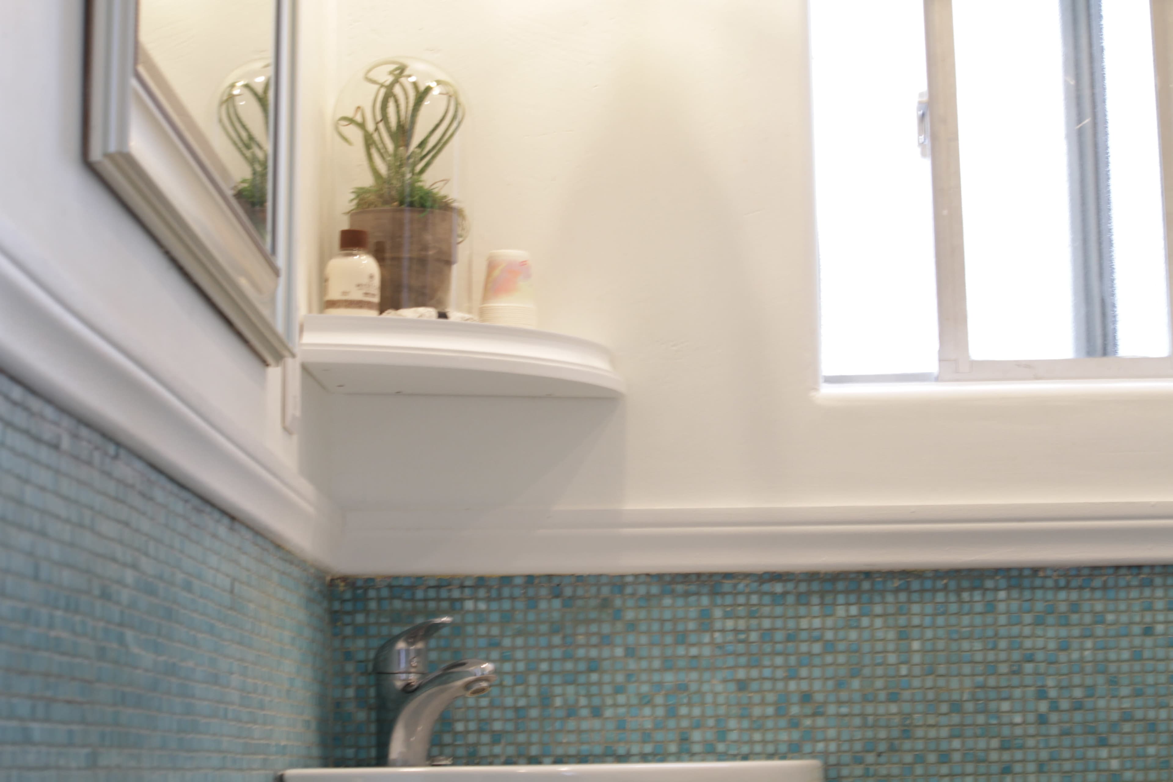 The image shows a bathroom with a sink, a small shelf containing toiletries, and a mirror above the sink, all set against a tiled blue wall.
