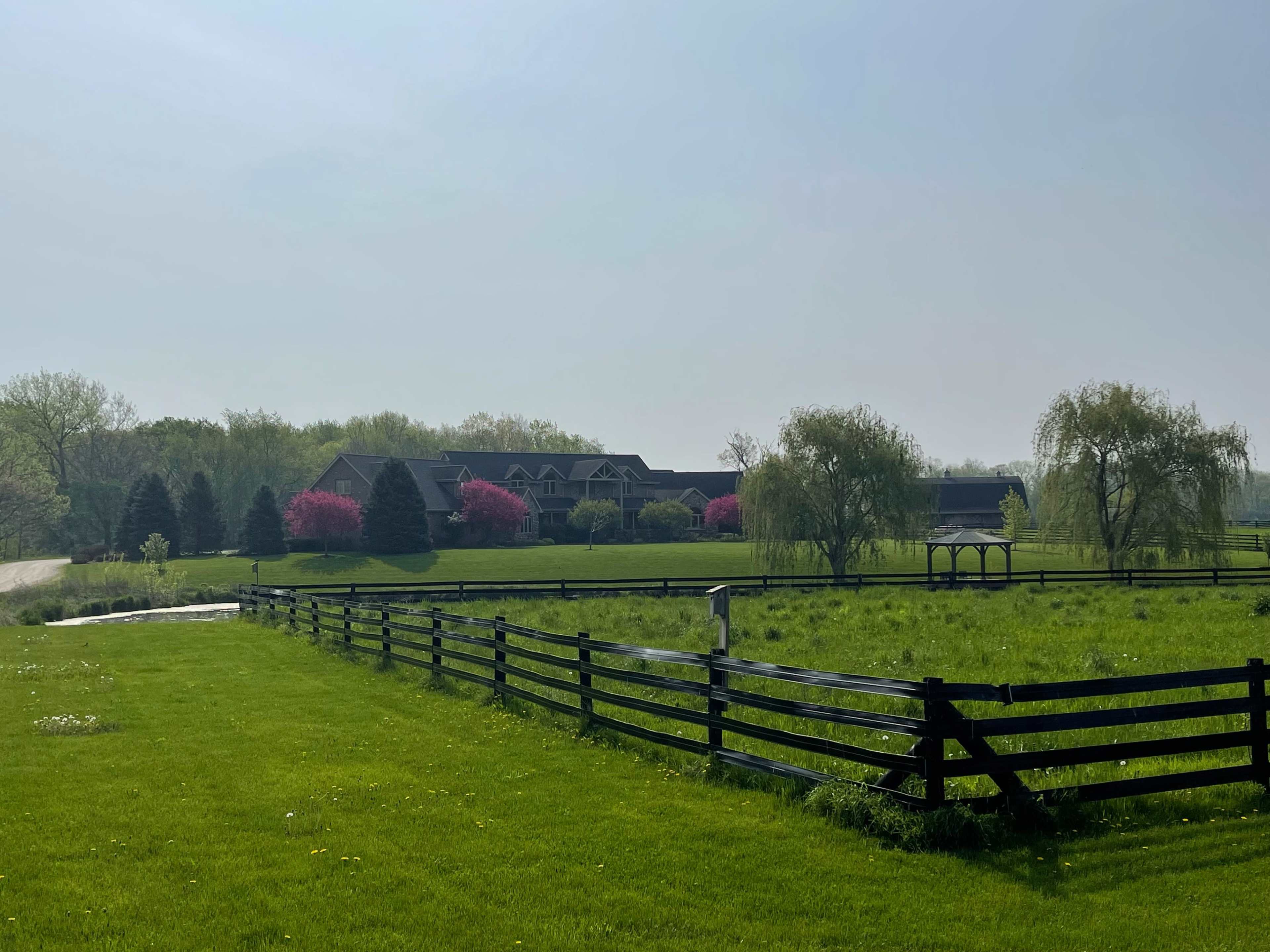A large house surrounded by trees and flowering plants is visible behind a black wooden fence in a green field.