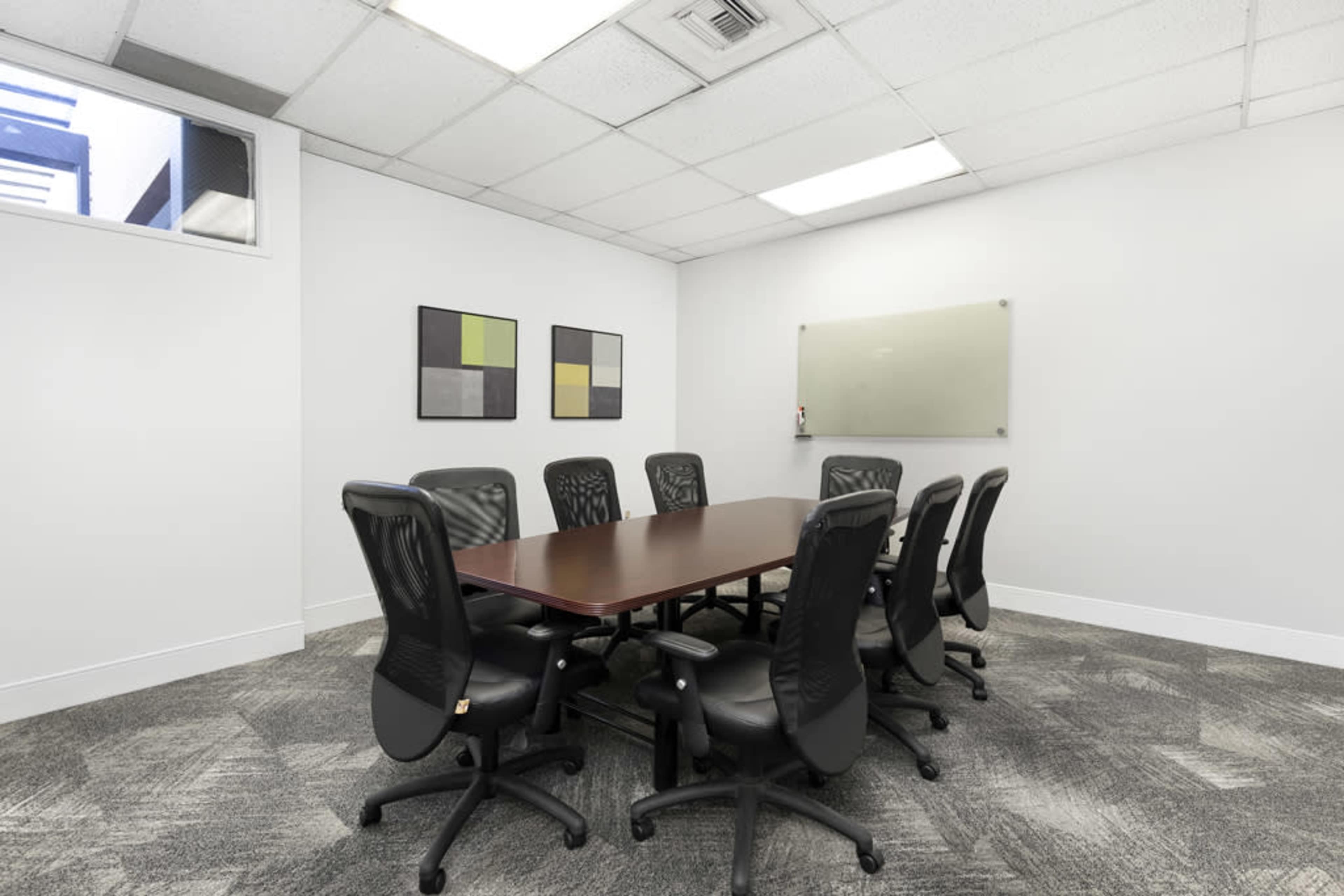 The image shows a conference room with a large rectangular table and eight black office chairs, set against white walls and minimal decorations.