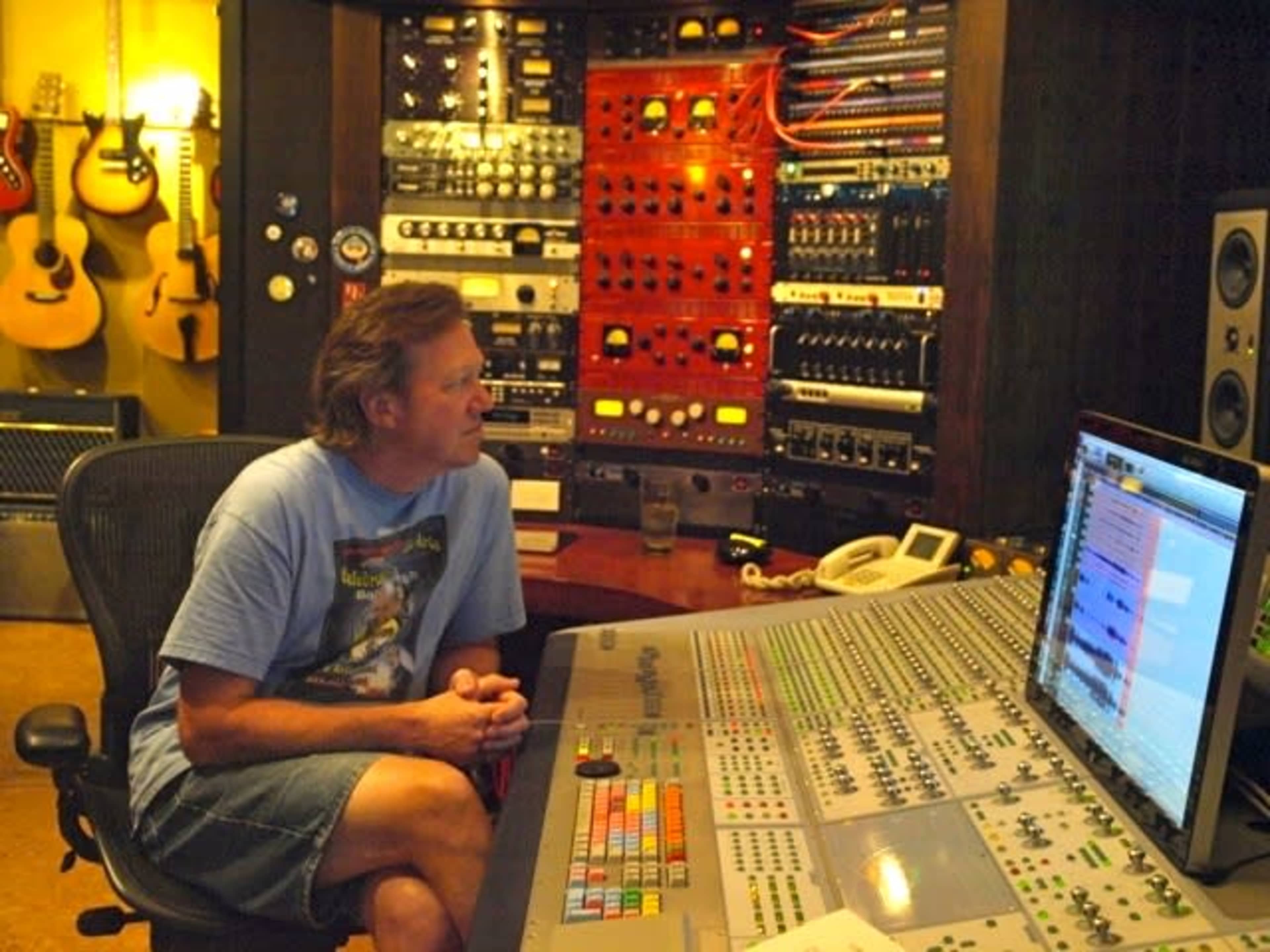 A man sits in front of a large audio mixing console in a recording studio, surrounded by various musical instruments and equipment.