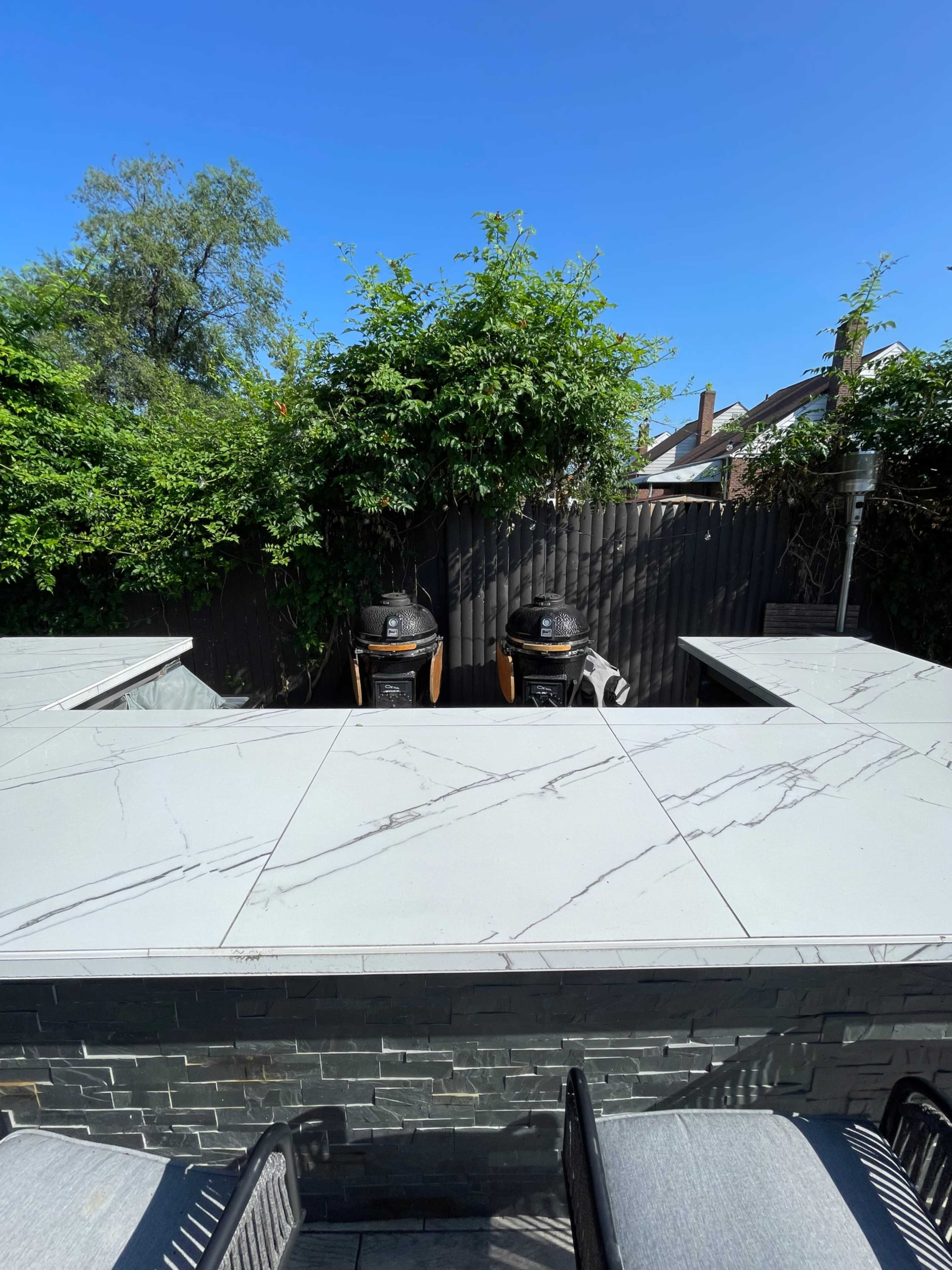 The image shows a modern outdoor kitchen area with two grills set against a backdrop of lush greenery and a clear blue sky.