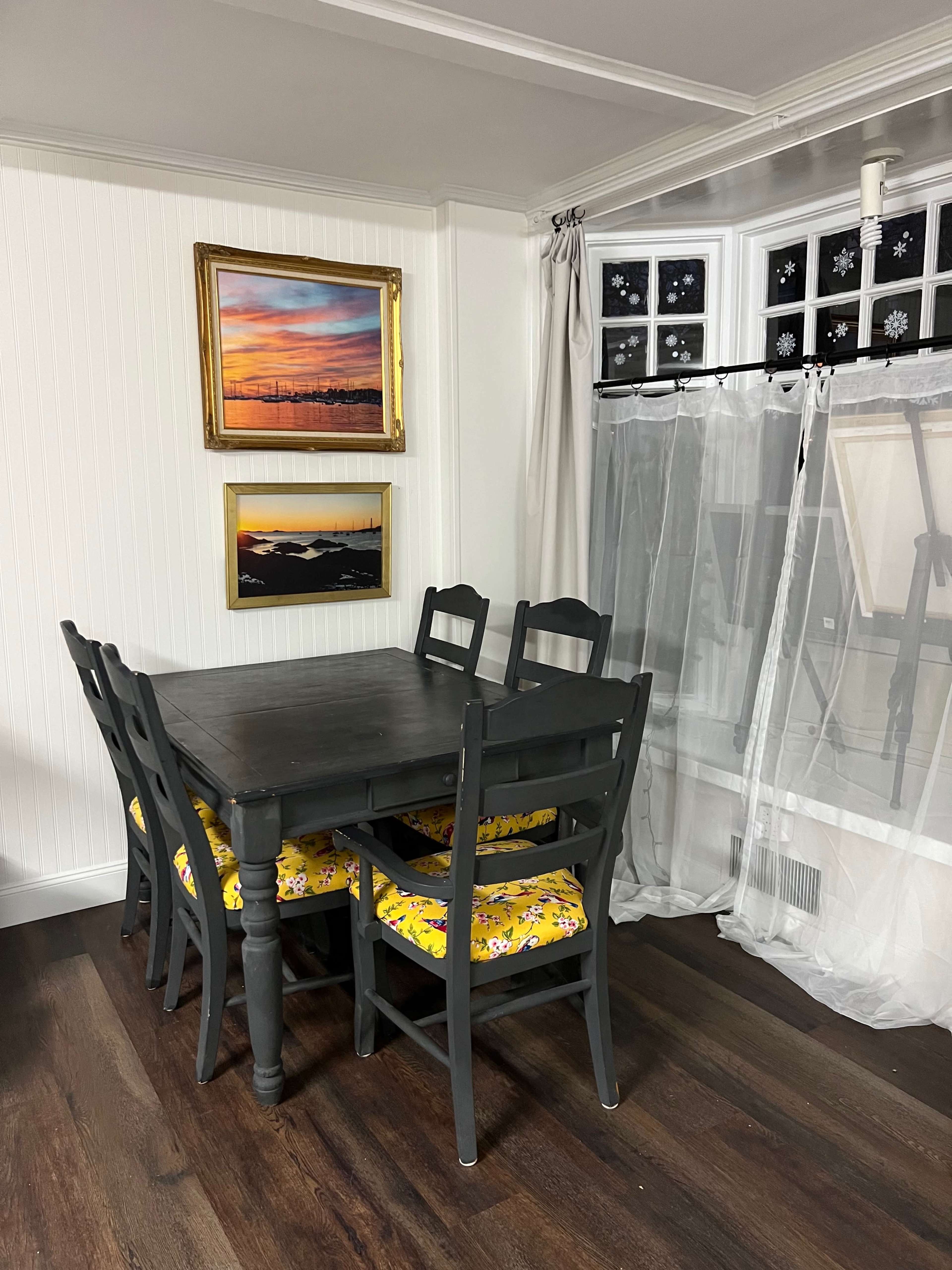 A black wooden dining table with six chairs, each featuring yellow floral cushions, is set against a white wall adorned with framed paintings and a window covered with sheer curtains.