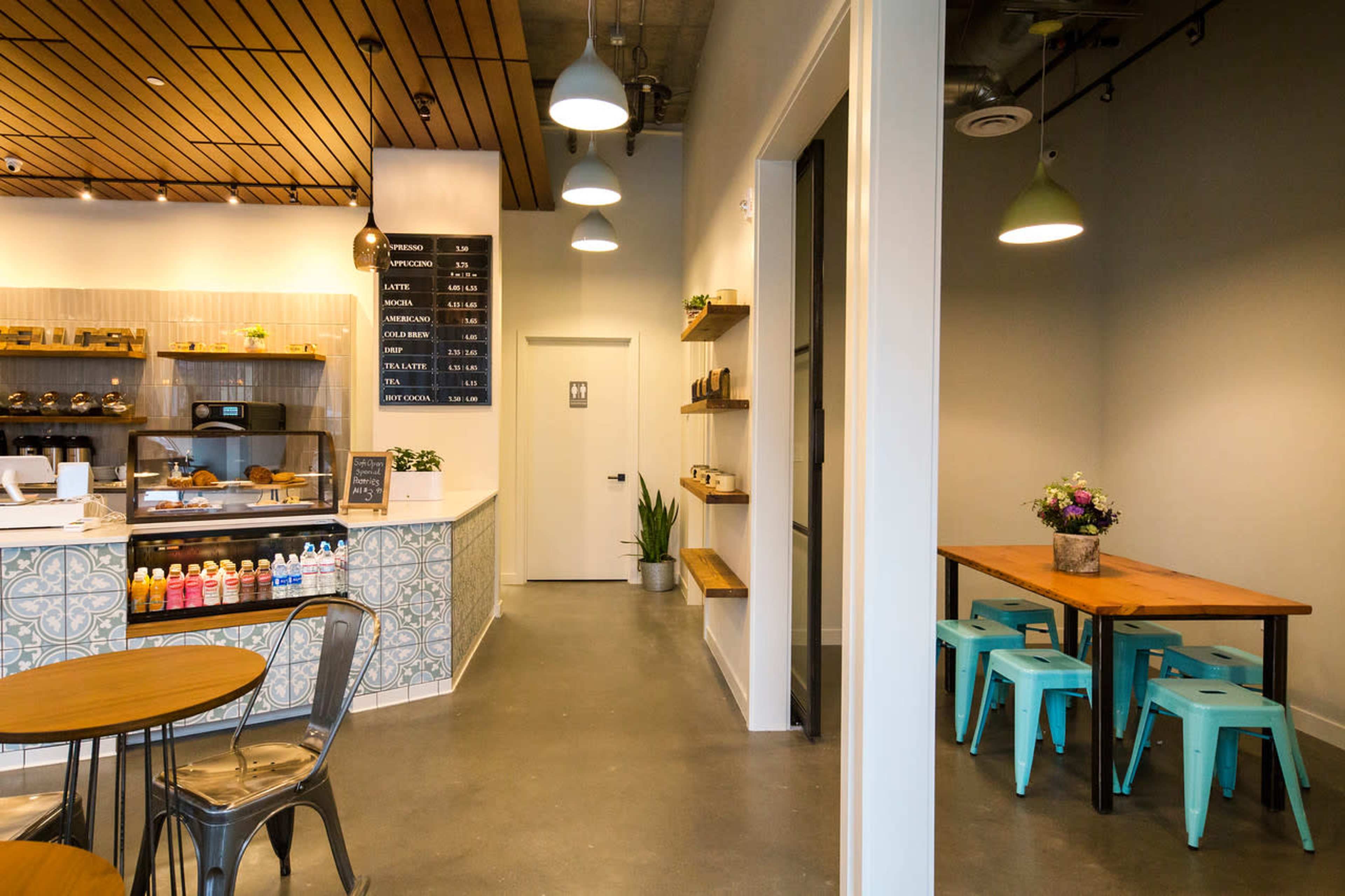 A modern café interior features a beverage counter, a menu board, and seating areas with wooden tables and teal chairs.