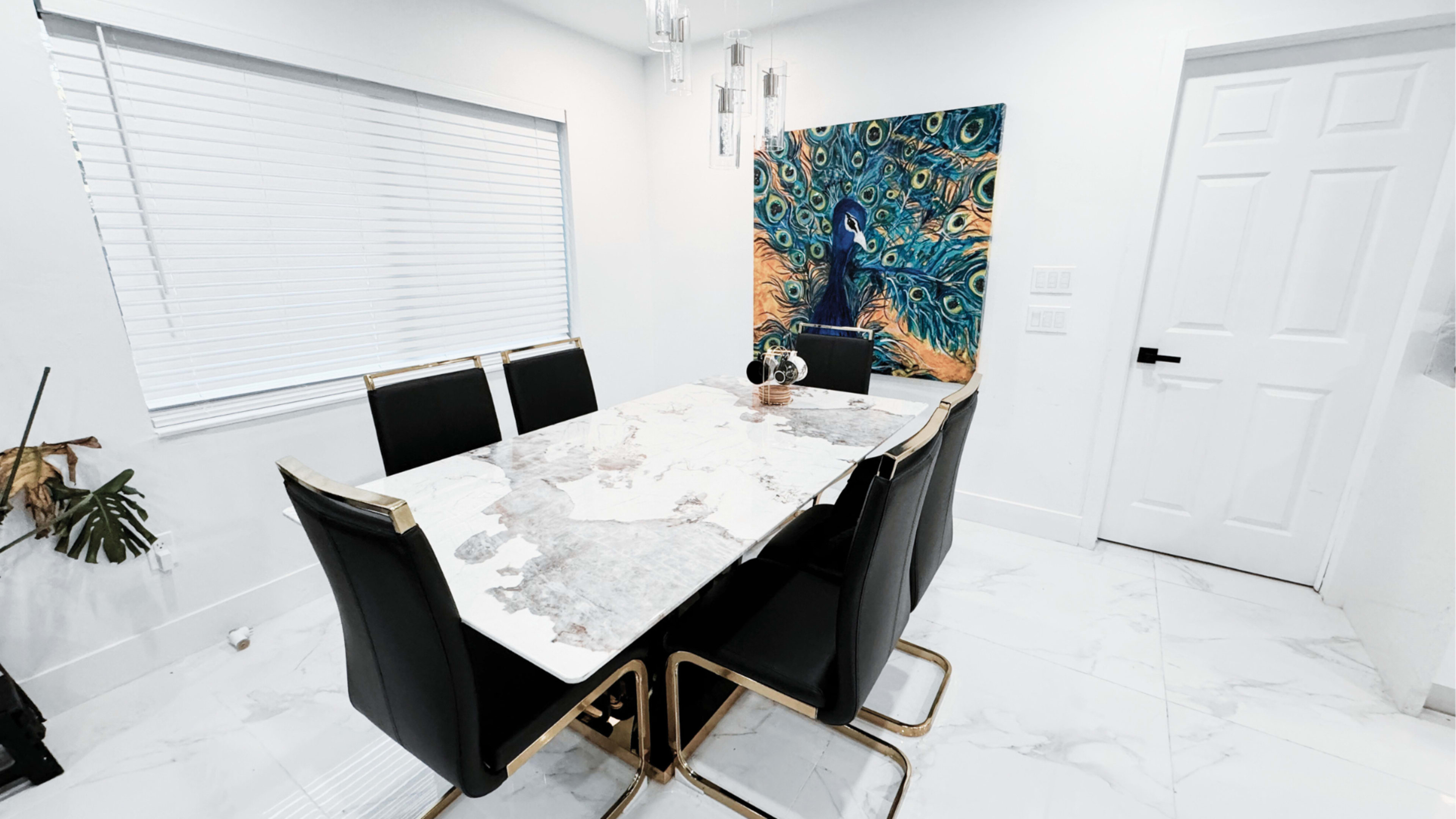 A dining room features a marble table surrounded by six black chairs, with a peacock painting on the wall and a door in the background.