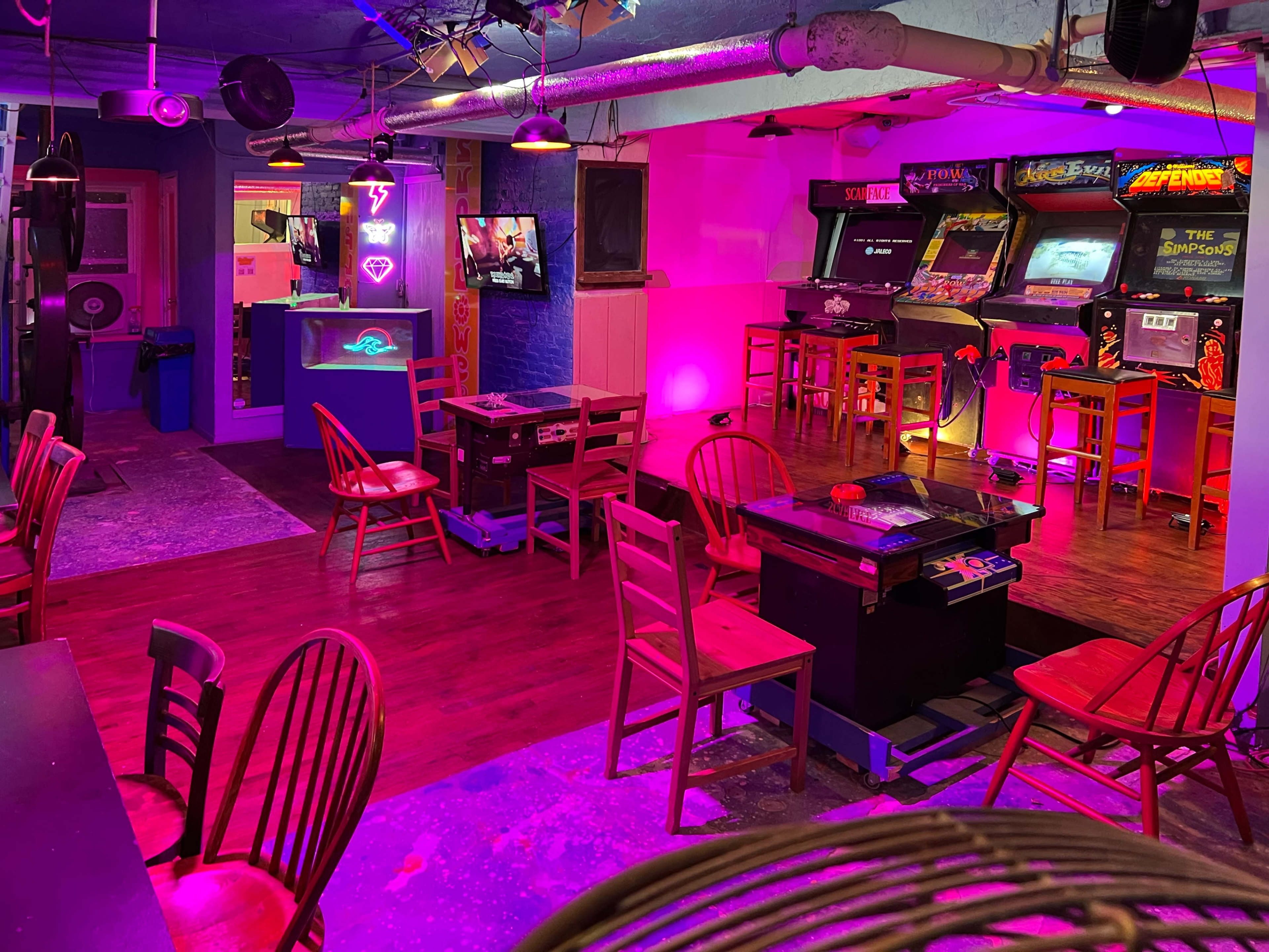 A dimly lit arcade room with neon lighting, featuring several arcade machines, empty chairs, and a bar area.