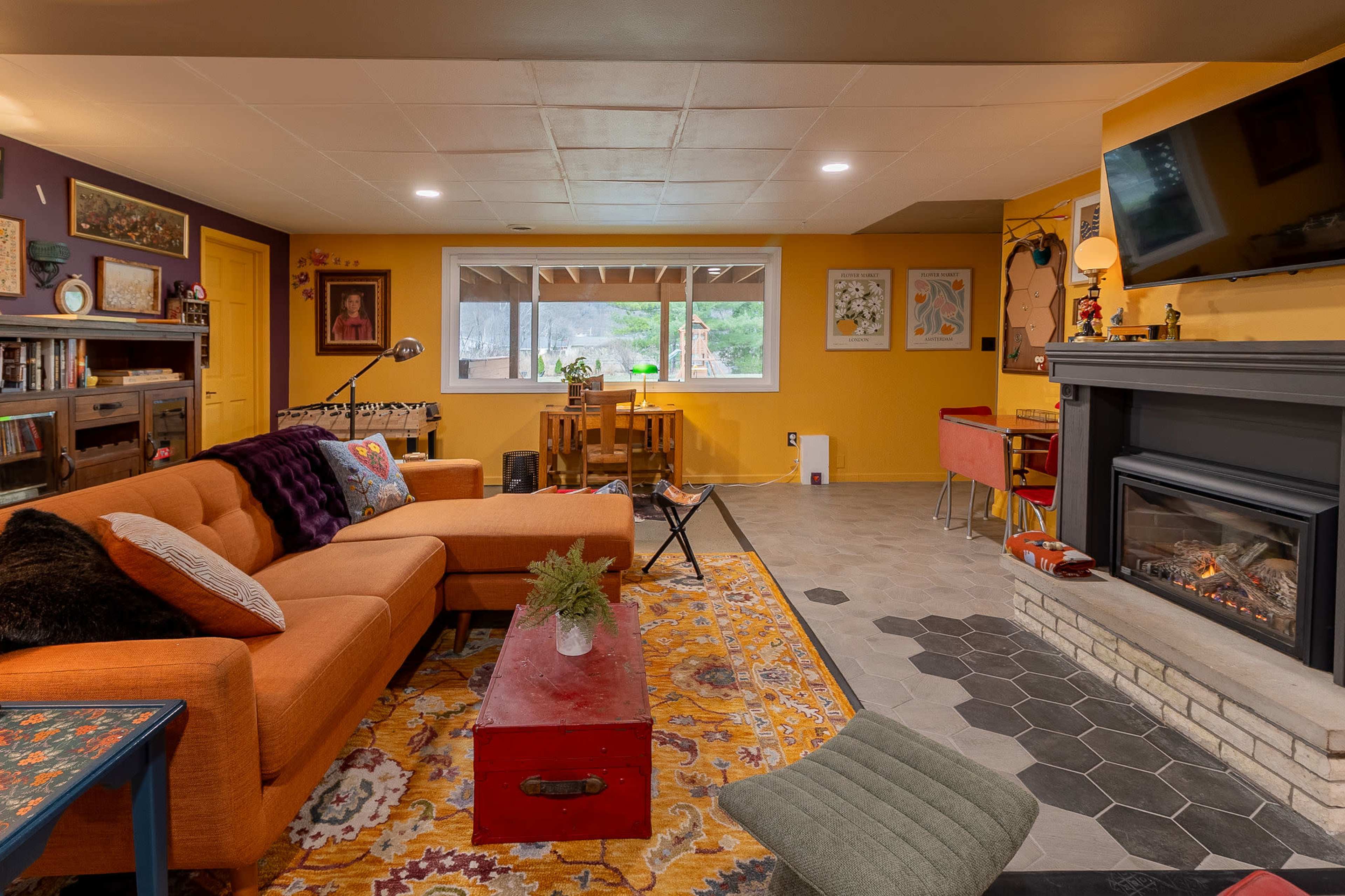 The image shows a cozy living room featuring a sectional sofa, a decorative rug, a fireplace, and a mix of colorful wall decor.