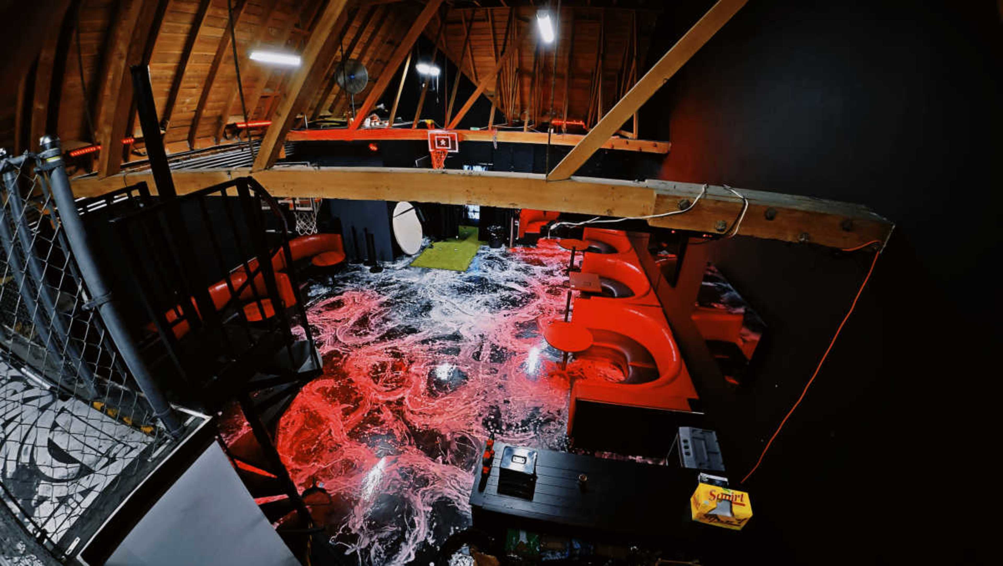 The image shows a dimly lit room with a black ceiling and wooden beams, featuring a striking red and white marbled floor and various red furnishings.