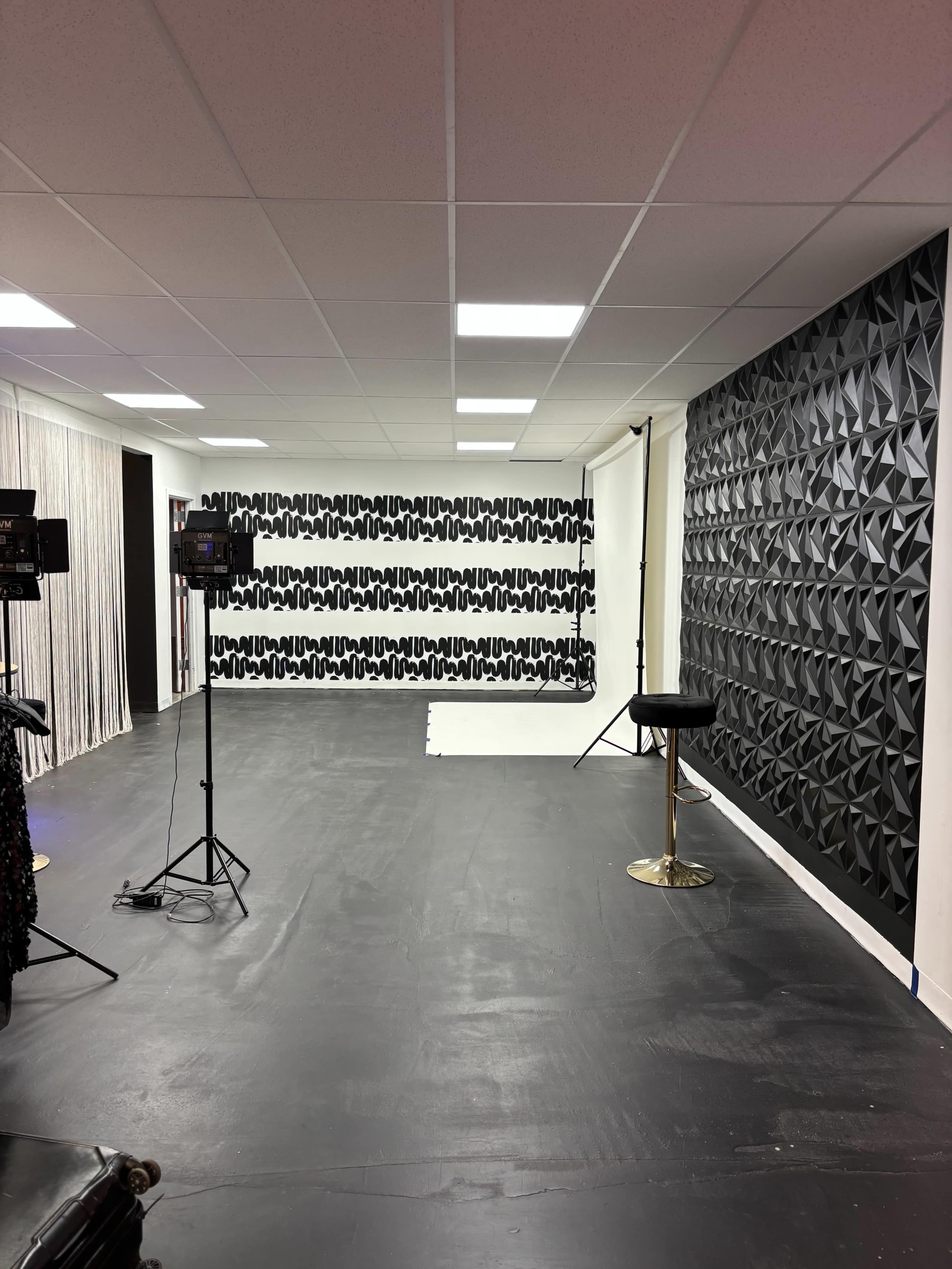 The image shows a spacious studio with black flooring, a textured black wall, and a patterned backdrop, equipped with lighting stands.