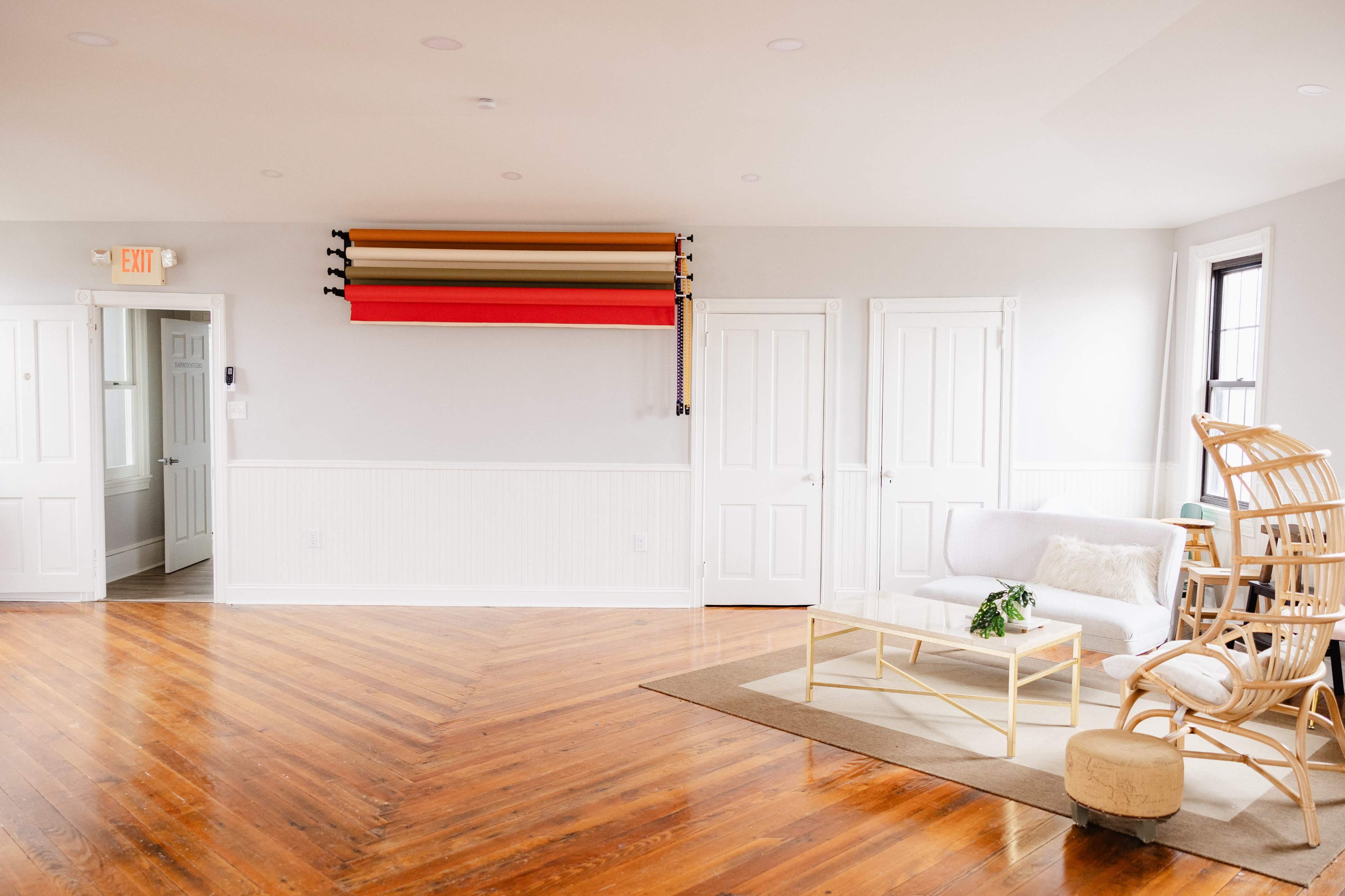 A bright, open room with hardwood floors, a white sofa, a rattan chair, and several large rolls of fabric mounted on the wall.