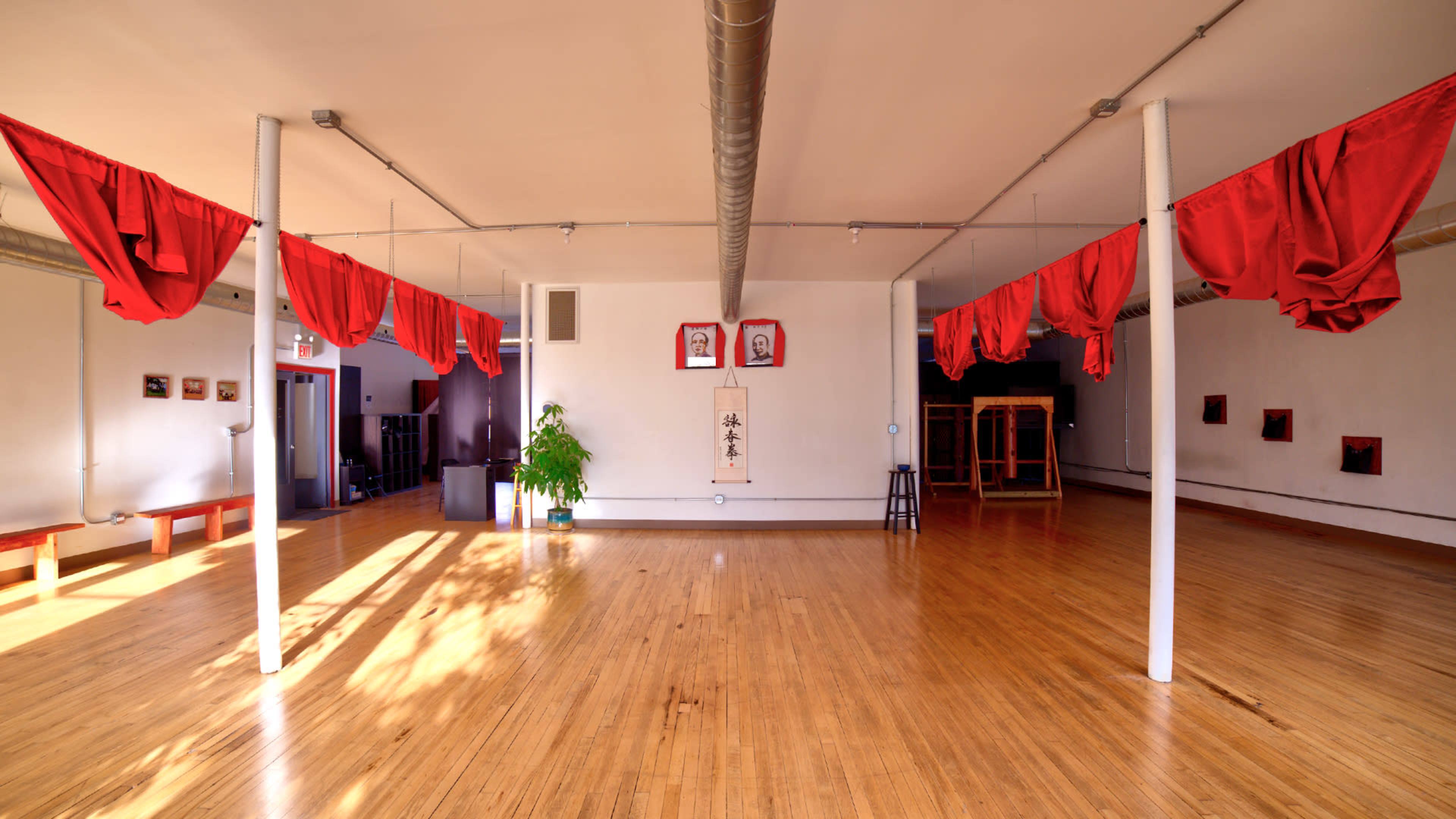 A spacious room with wooden flooring, red fabric draped from the ceiling, and a few pieces of art on the wall.