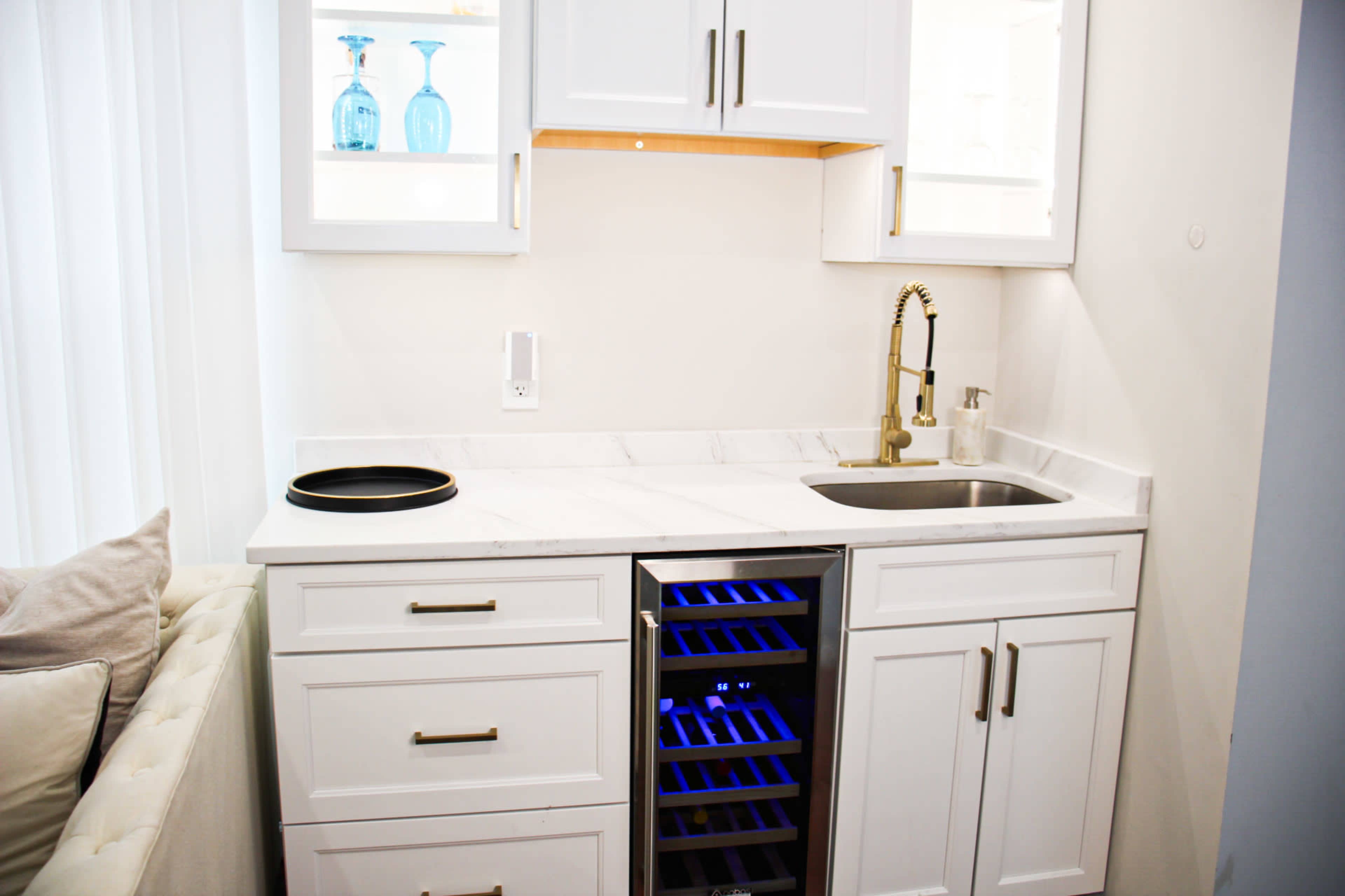 A modern kitchenette with white cabinets, a marble countertop, a sink, and a wine refrigerator with blue lighting.