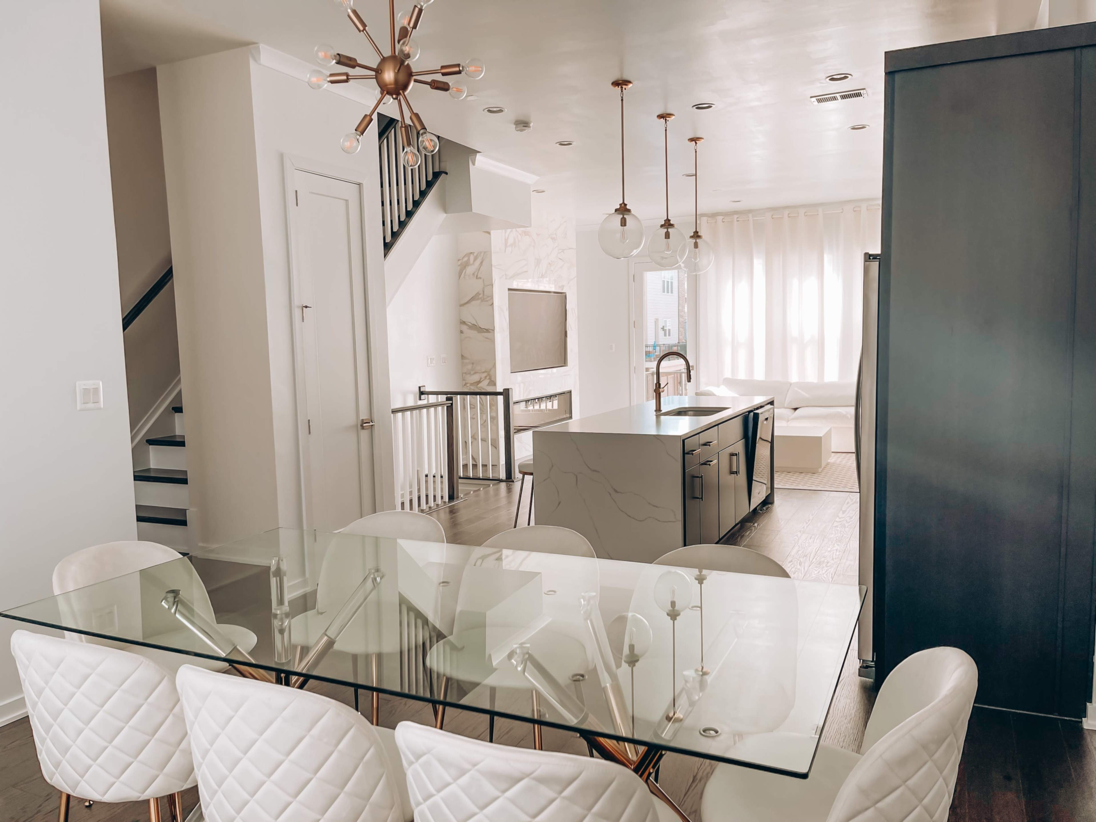 A modern dining area with a glass table and white chairs, adjacent to a kitchen island, and a staircase leading to an upper level.