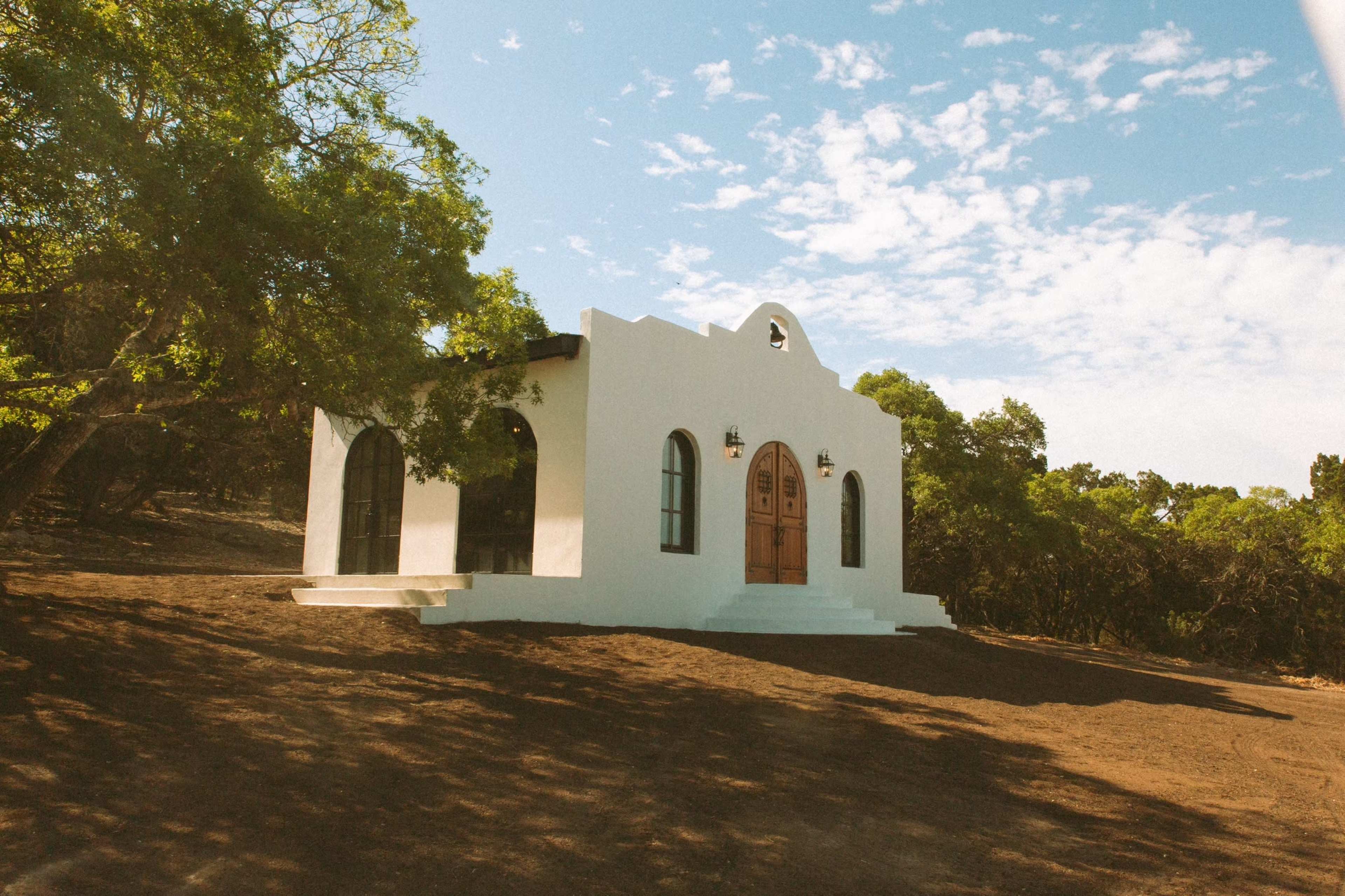 A white, stucco building with arched windows and wooden double doors sits on a hillside surrounded by trees under a blue sky.