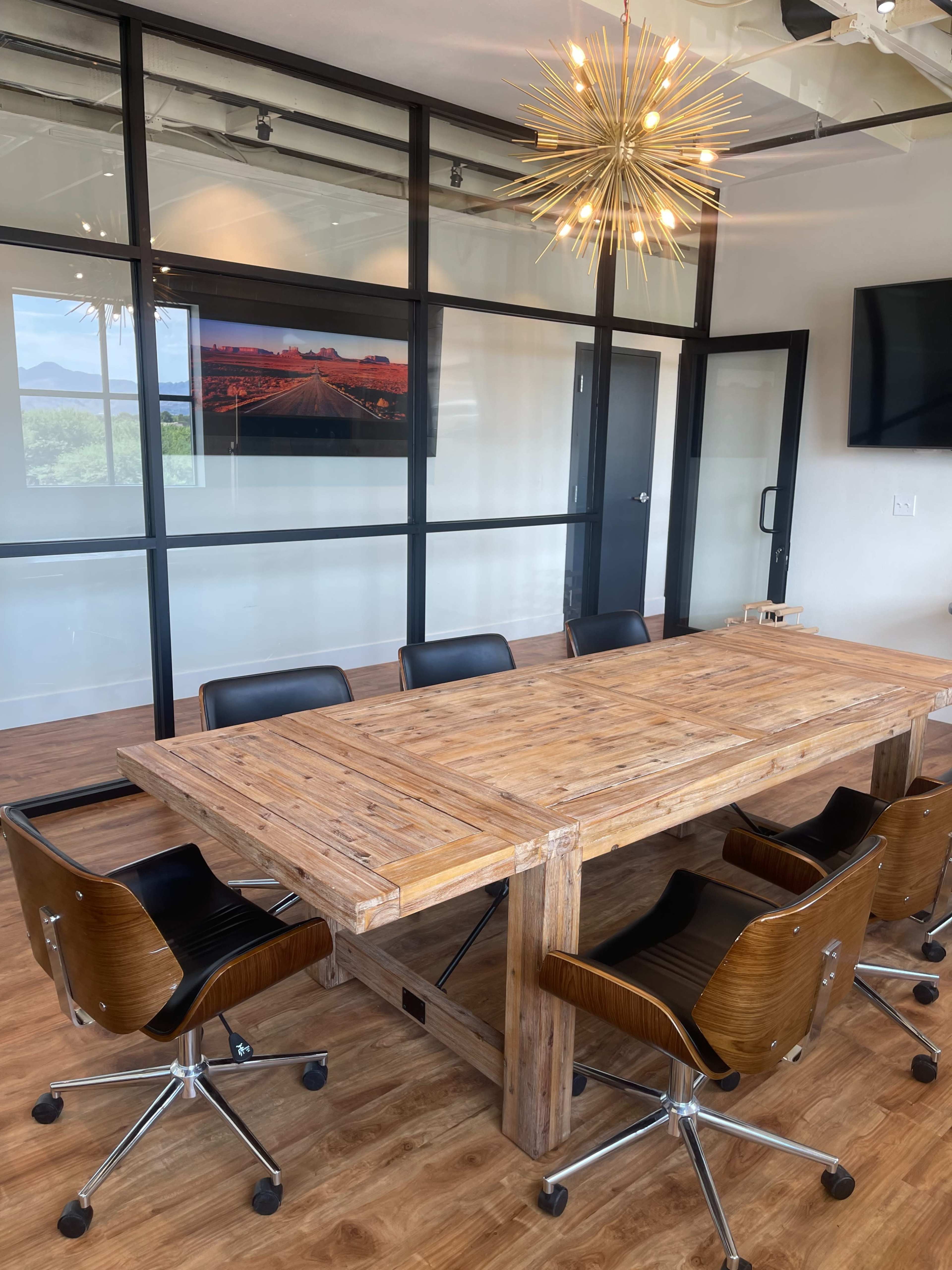 A conference room features a large wooden table surrounded by black rolling chairs, with glass walls and a decorative light fixture above.