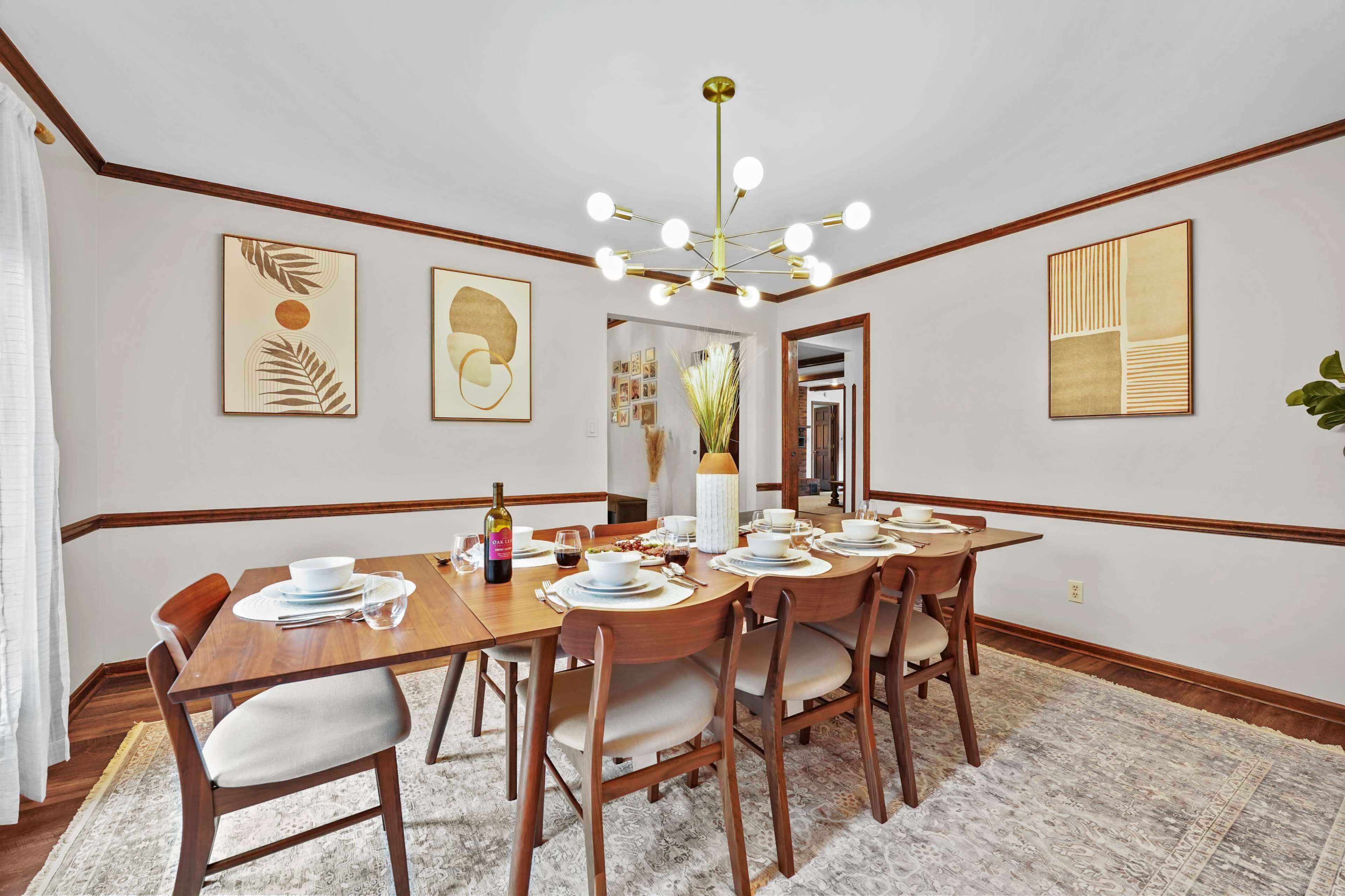 A dining room features a wooden table set for a meal, surrounded by chairs, with decorative wall art and a modern light fixture.