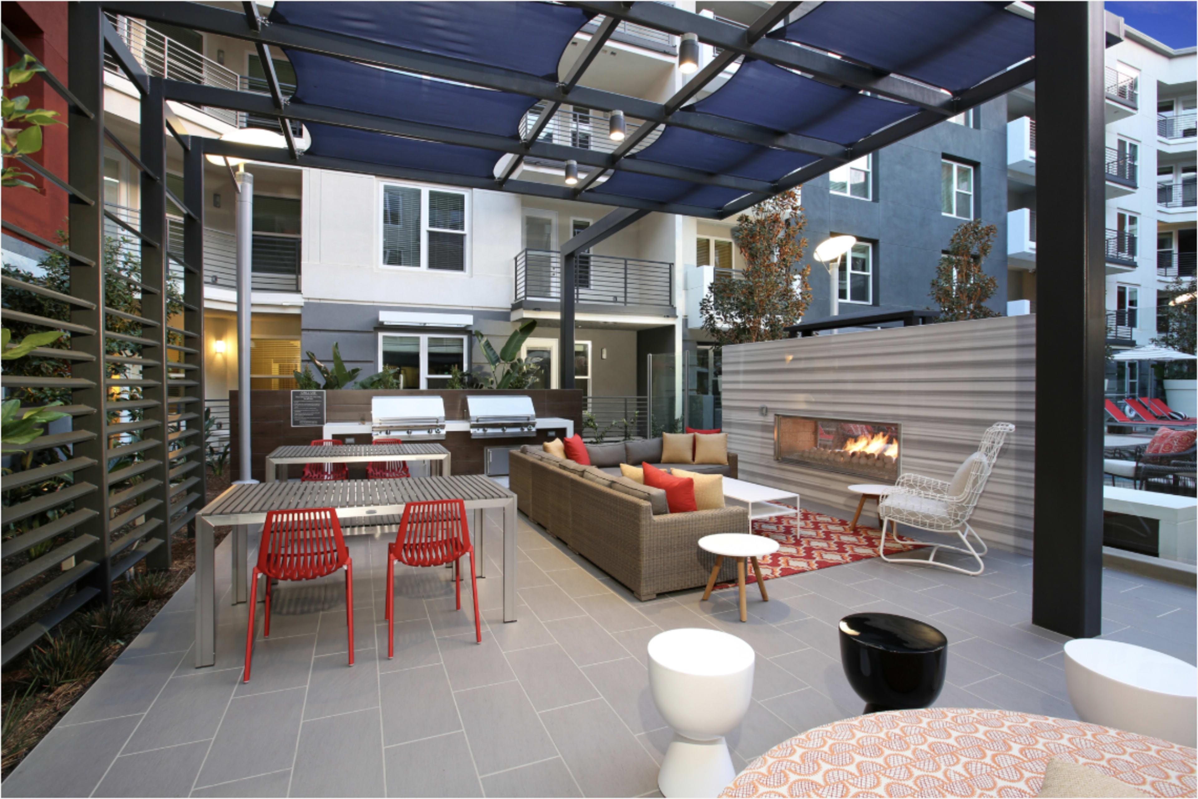 The image shows an outdoor lounge area with seating, a fire pit, a dining table, and a shaded pergola, situated next to modern apartment buildings.