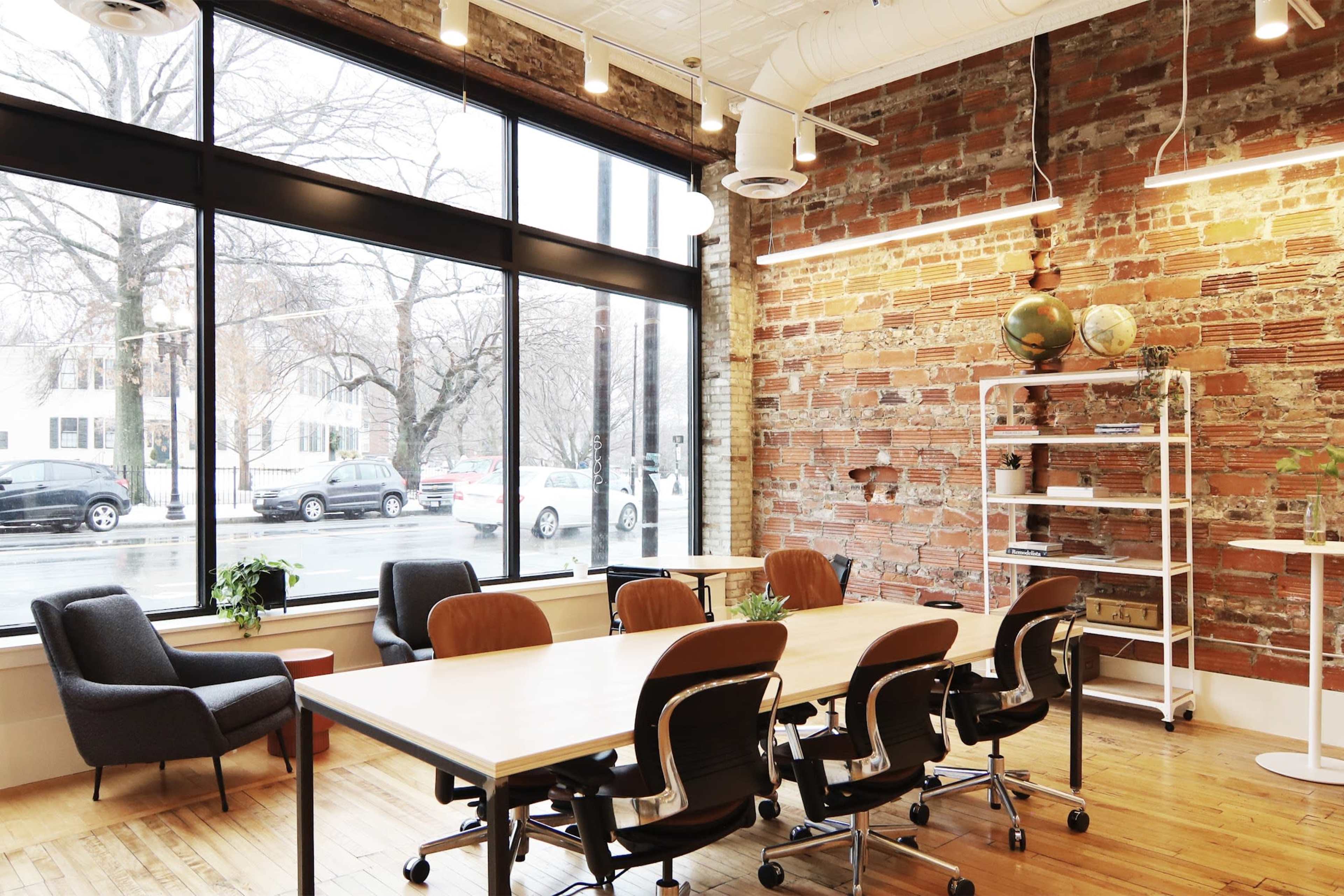 A modern conference room features a wooden table surrounded by black office chairs, large windows revealing a street view, and exposed brick walls.