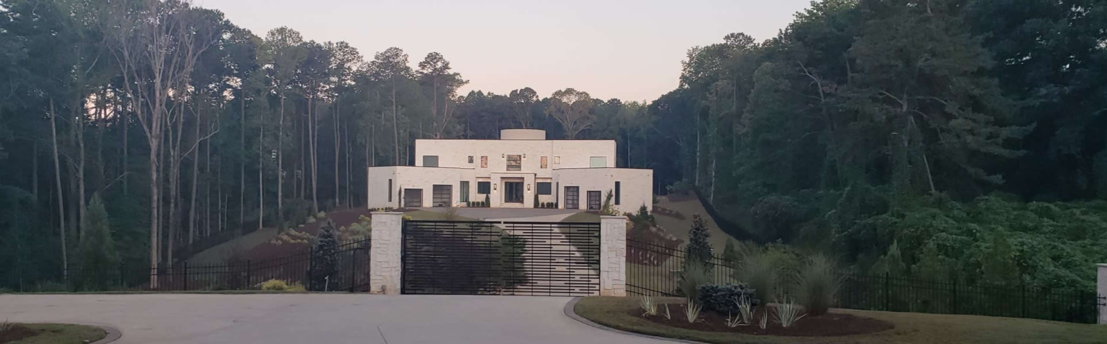 A modern, white multi-story house sits at the end of a driveway surrounded by tall trees and lush greenery.